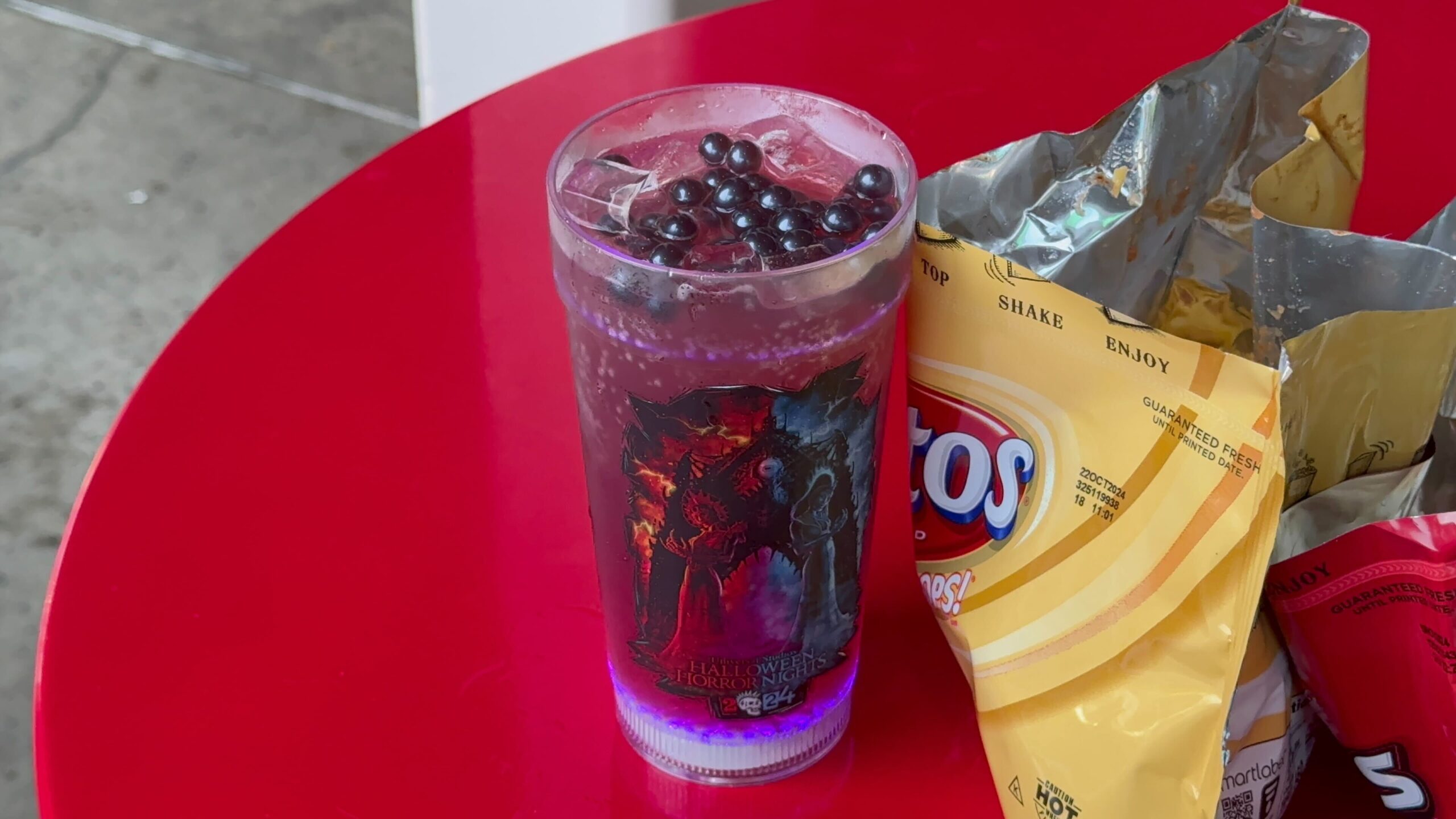 A glass of dark bubble tea with tapioca pearls on top, placed next to an open bag of yellow Lay's chips on a red table. Perfect for a quick snack as you get ready for Halloween Horror Nights 33!
