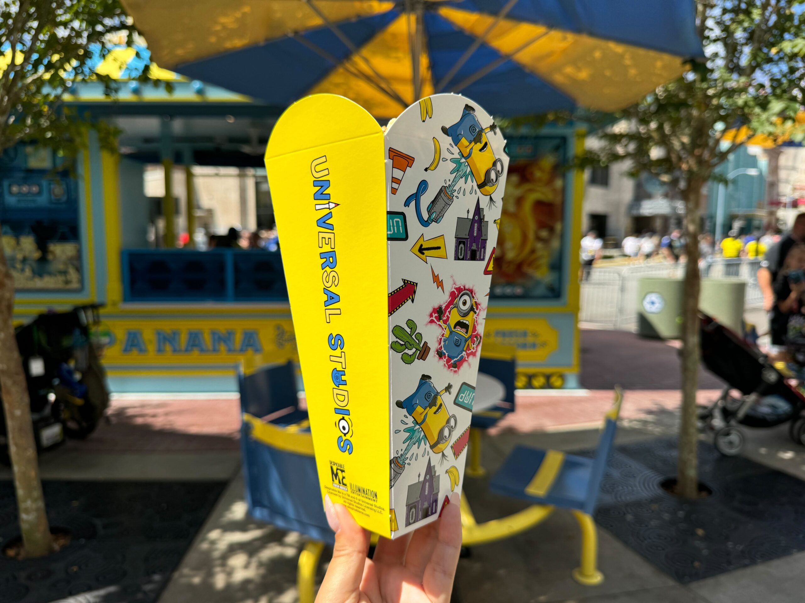 Close-up of a person holding a souvenir popcorn box with Universal Studios branding in a theme park, with an outdoor seating area and a concession stand visible in the background.