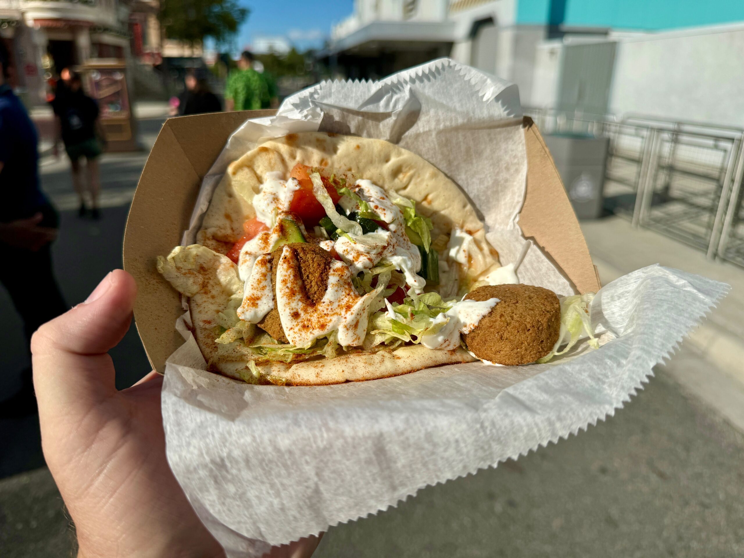 A hand holding a cardboard container with a falafel pita wrap filled with falafel balls, lettuce, tomatoes, and drizzled with a creamy sauce, against an outdoor urban backdrop. Perfect for those nights when you're exploring the city or need something savory before heading to Halloween Horror Nights.