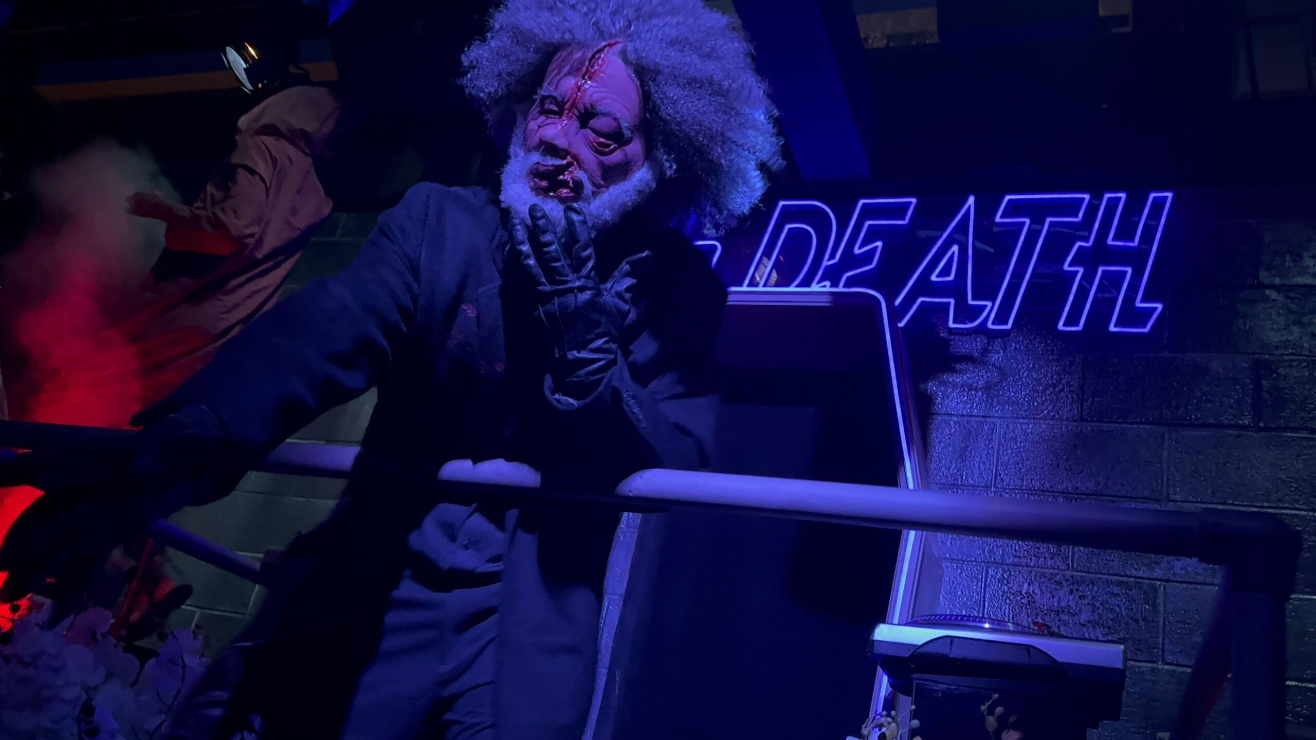 A person in a detailed monster costume with wild hair and a split face stands in a dark, neon-lit environment with the word "DEATH" illuminated behind them.