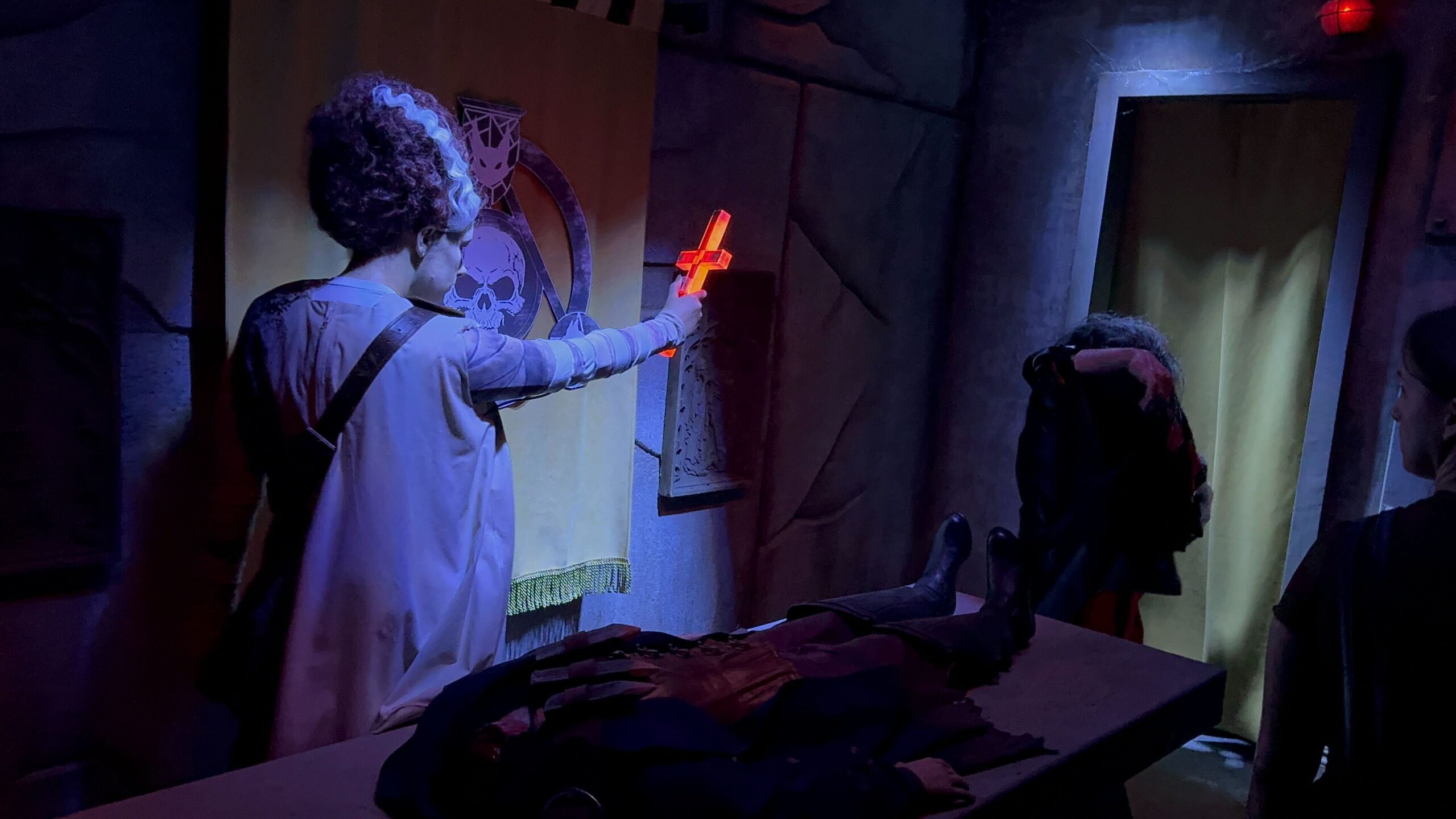 A person in a costume with a glowing red cross faces another person in a dark room with a skull banner.