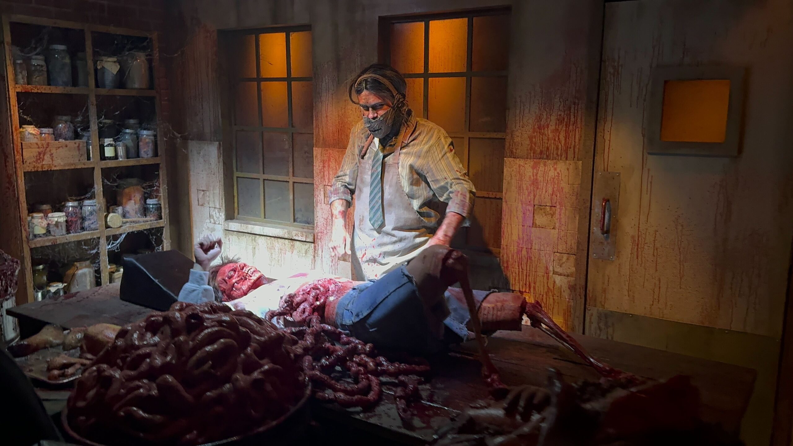 A person appears to be conducting surgery or an autopsy on a human body in a dimly lit, blood-stained room with scattered entrails, reminiscent of the grisly scenes from Texas Chainsaw Massacre.