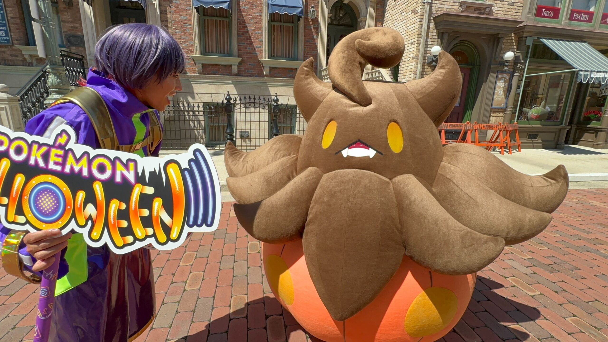 A person in a colorful outfit interacts with a large, plush Pokémon character in a pumpkin-like costume on a brick street. The person holds a sign that partially reads "POKÉMON GO.