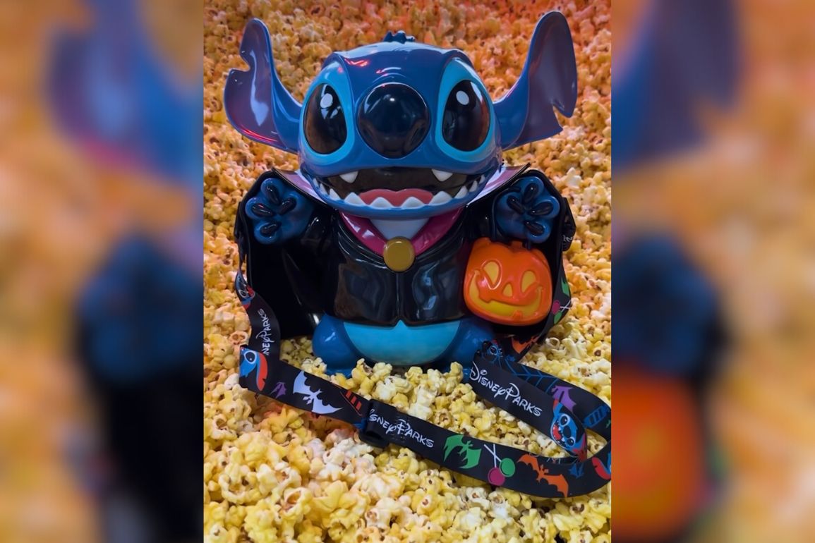 A Vampire Stitch figurine holds a jack-o'-lantern bucket, surrounded by popcorn. He sports a lanyard with Halloween designs, making it the perfect Disneyland popcorn bucket souvenir.