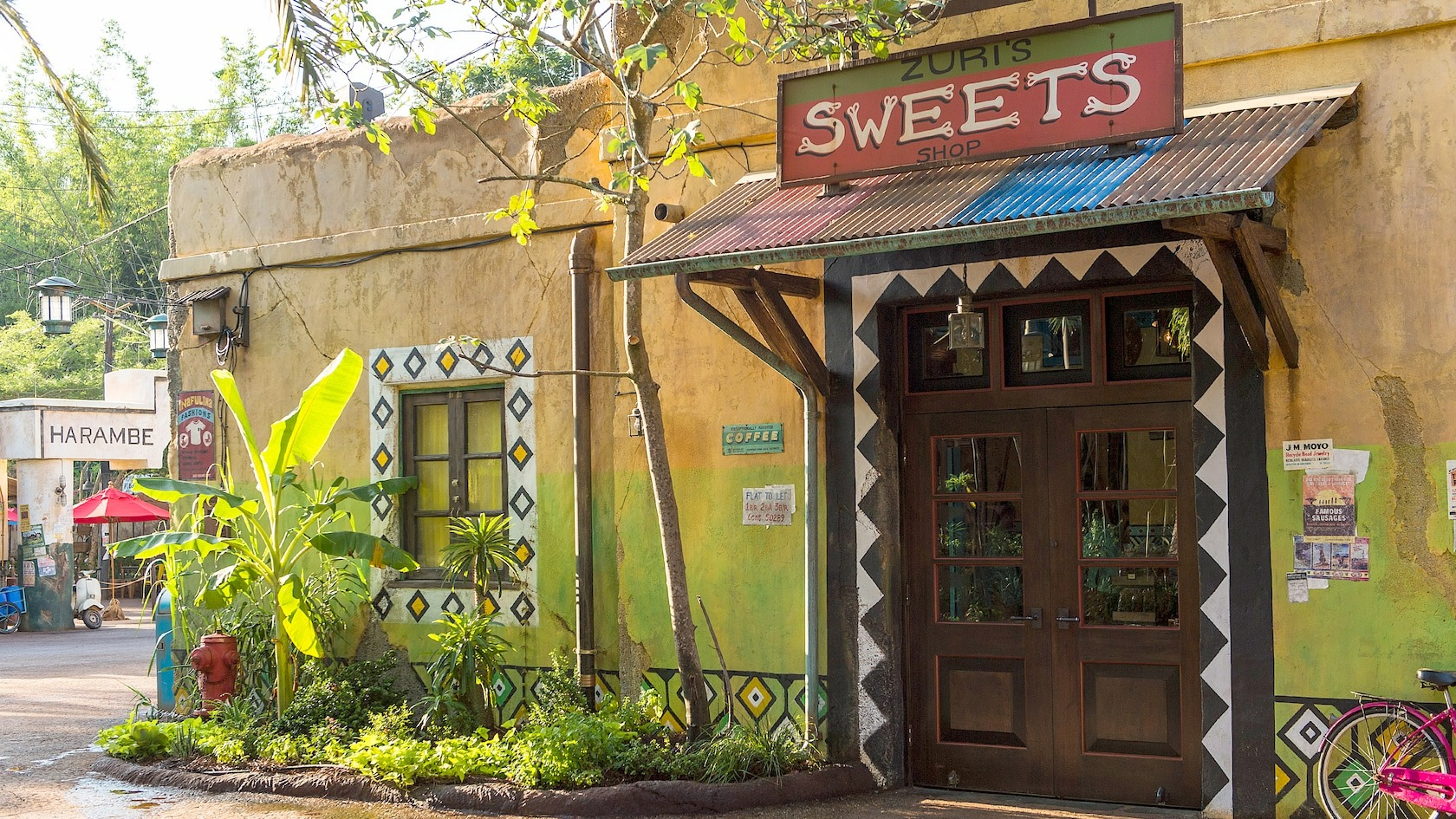 A quaint shop named "Zuri's Sweets Shop" with a colorful sign, wooden door, and decorative walls. Greenery and a fire hydrant are visible along the sidewalk. The area appears bright and sunny, inviting visitors to try their famous Zebra Cupcakes inspired by Disney's Animal Kingdom.