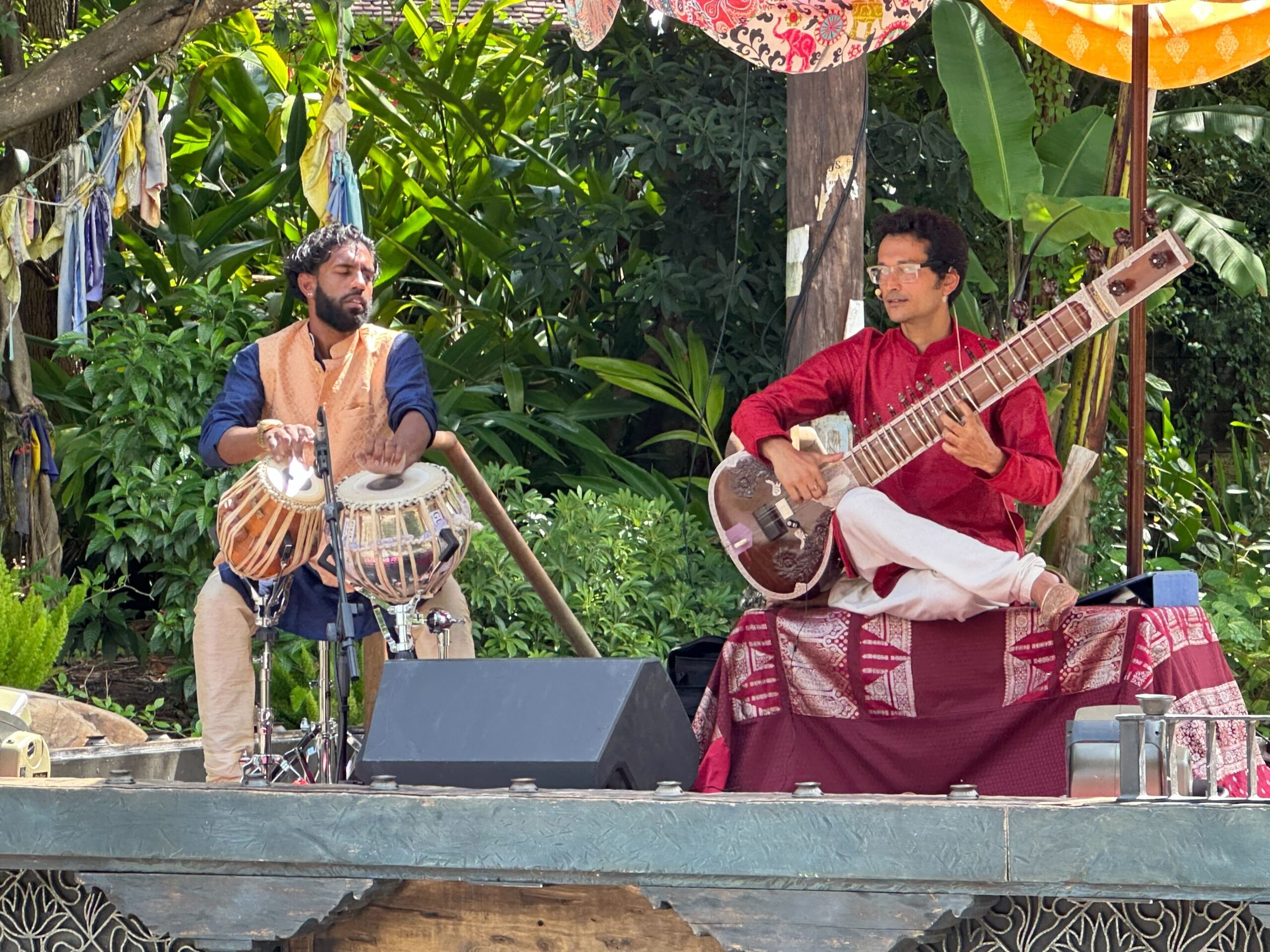 Two musicians perform outdoors. One plays a sitar while seated on a platform draped with fabric. The other plays a pair of tabla drums. There is lush greenery in the background.