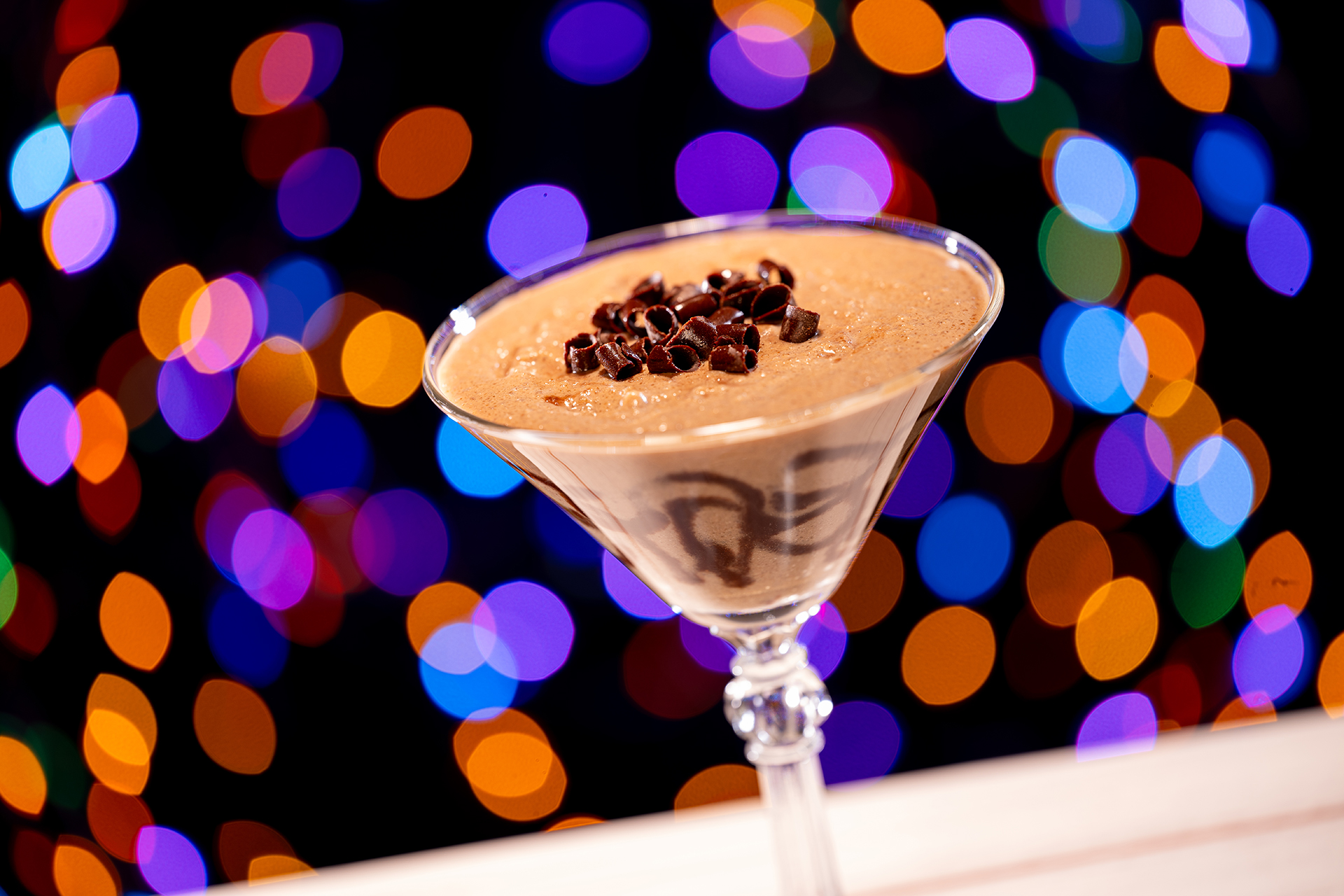 A martini glass filled with a brown dessert, topped with chocolate shavings, glistens against a backdrop of colorful, out-of-focus lights at Disney Jollywood Nights 2024.