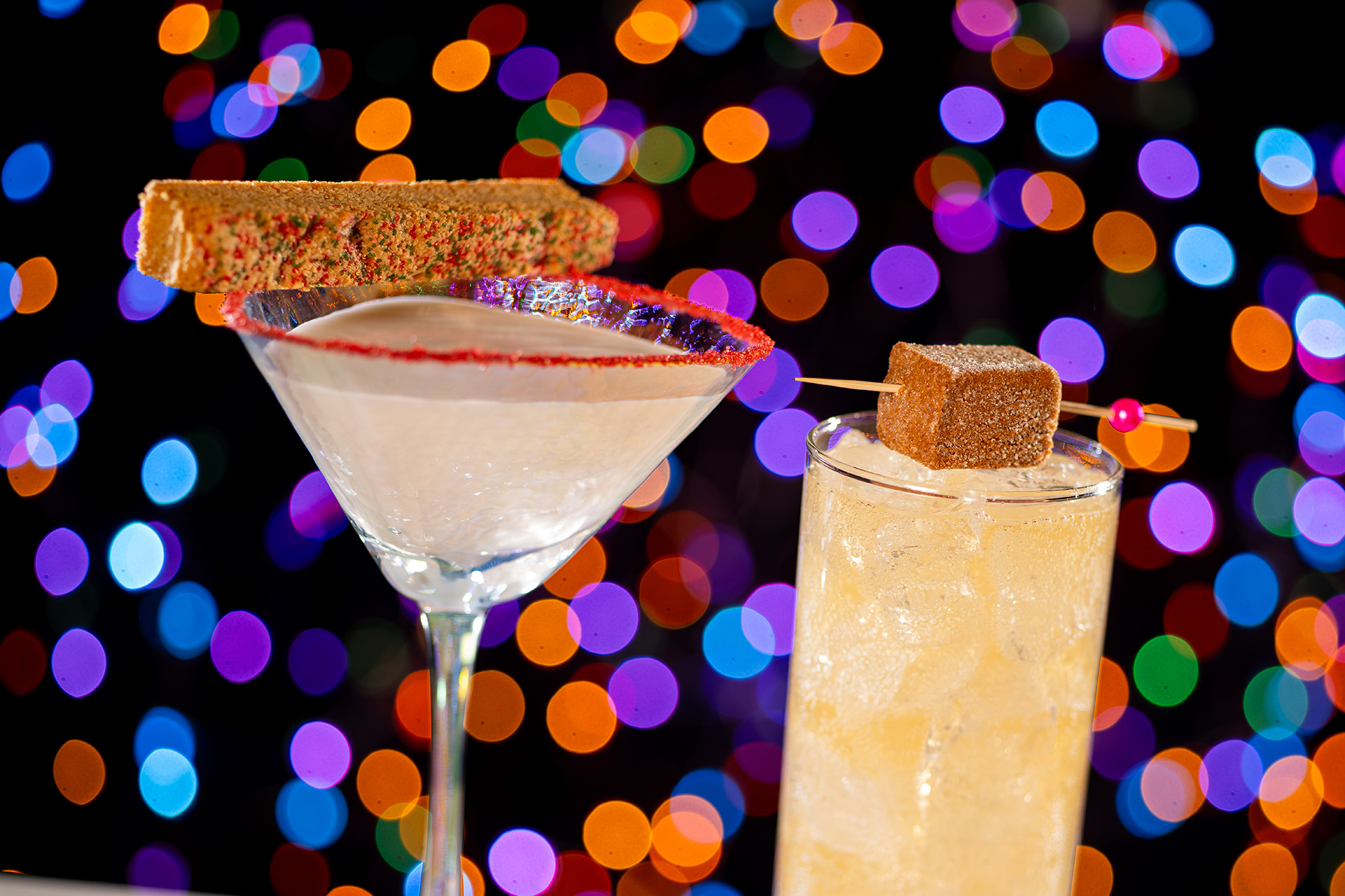 Two festive cocktails, one in a martini glass with a biscotti garnish and another in a highball glass with an ice cube and a small garnish, set against colorful blurred lights, perfect for enjoying during Disney Jollywood Nights' food and beverages offerings.