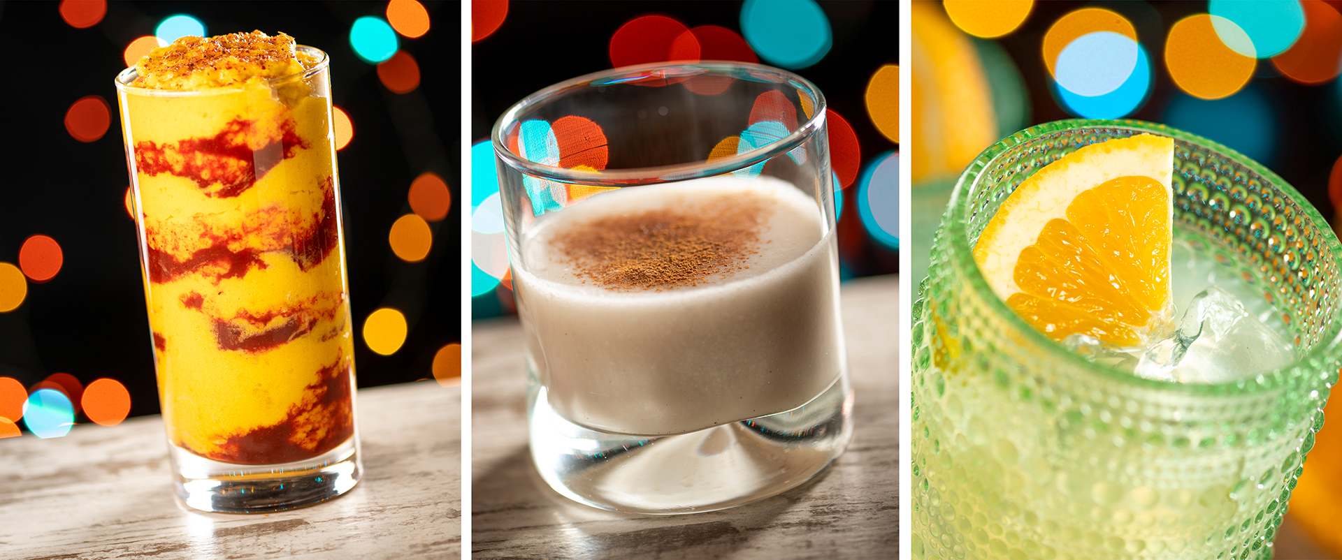 At Disney Jollywood Nights 2024, enjoy three exquisite cocktails: a tall glass with a yellow and red layered drink, a short glass with a creamy beverage and a hint of cinnamon, and a green glass with a clear drink garnished with an orange slice. Savor the magic in each sip!