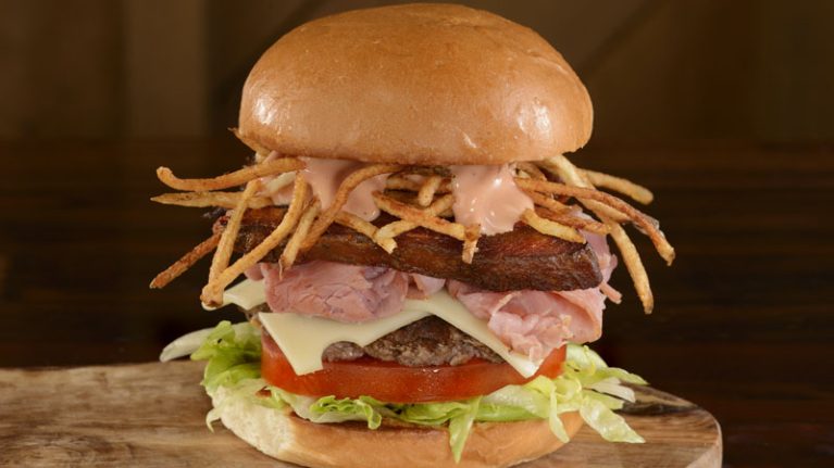 A large burger with lettuce, tomato, cheese, a beef patty, ham, crispy fried shoestring potatoes, and sauce inside a bun sits enticingly on a wooden surface—perfect for celebrating Hispanic and Latin American Heritage Month with delicious food and beverages.