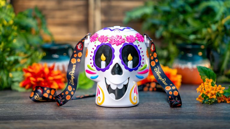 A Day of the Dead-themed skull mug with colorful decorations and a black strap featuring floral designs, set against a background of green foliage and orange flowers. Celebrate Hispanic and Latin American Heritage Month with this vibrant piece, perfect for enhancing your Food and Beverages collection.