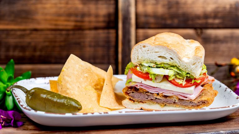 A plate containing a sandwich with layers of meats, cheese, tomatoes, and lettuce next to tortilla chips and a green chili pepper, reminiscent of the flavors celebrated during Hispanic and Latin American Heritage Month.