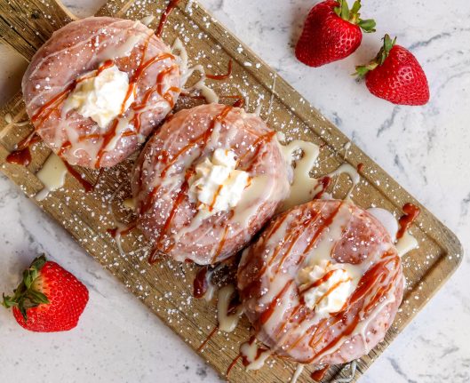 Three glazed strawberry donuts topped with whipped cream and various drizzles are arranged on a wooden board, with three whole strawberries placed around it. This delightful treat could easily be a hit at Walt Disney World during Hispanic and Latin American Heritage Month celebrations.