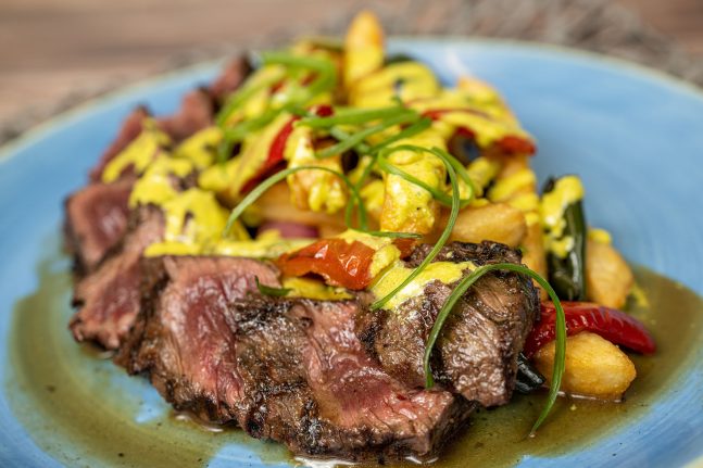 A plate of sliced grilled steak topped with green herbs, accompanied by roasted vegetables and drizzled with yellow sauce—a culinary delight celebrating Hispanic and Latin American Heritage Month at Disneyland and Walt Disney World.