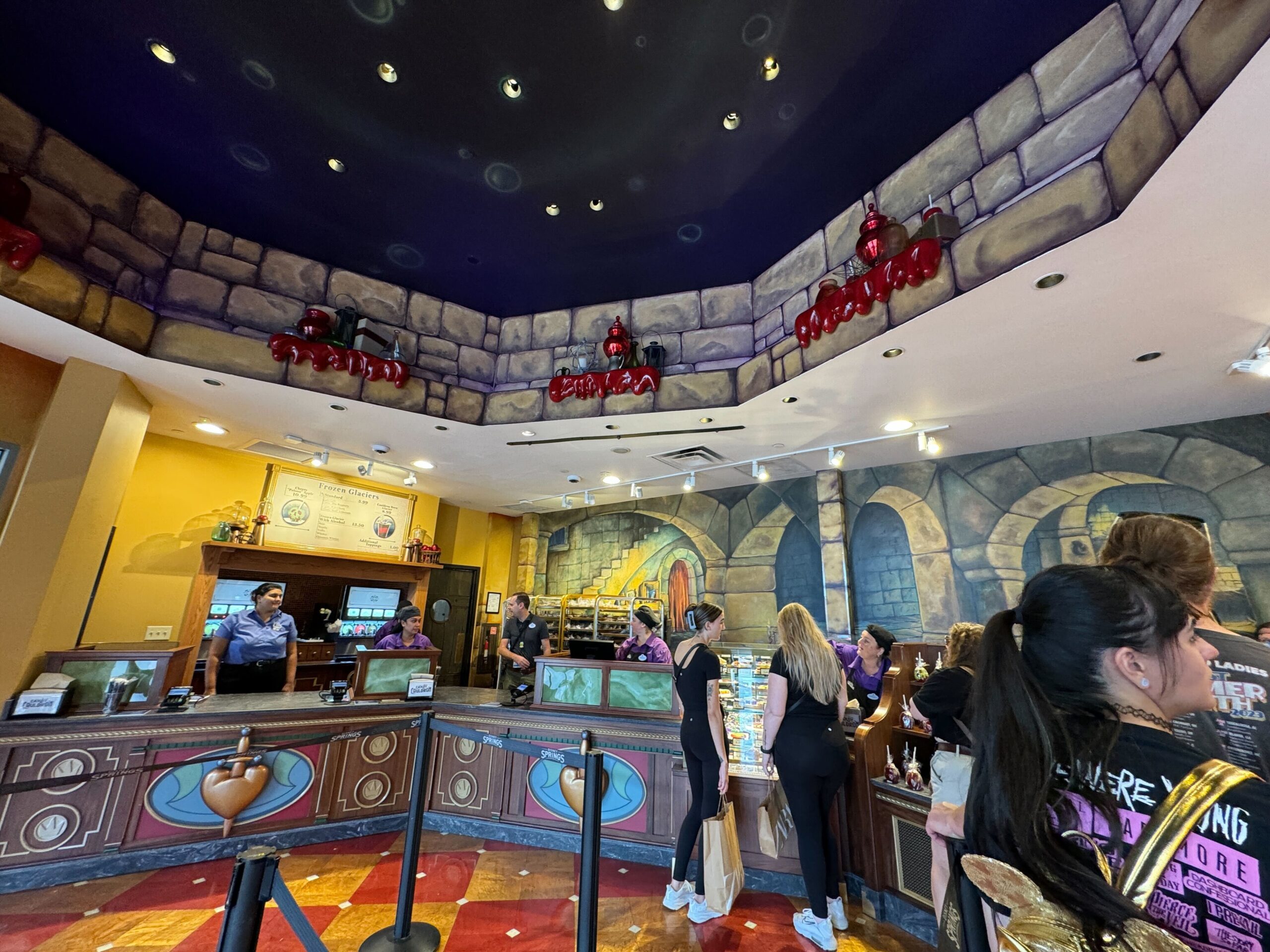 A themed indoor restaurant with a castle-like decor. Several employees are behind the counter, serving customers who are lined up. The ceiling has decorative chandeliers, and the walls mimic stonework.