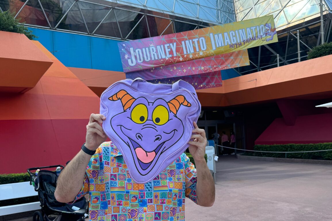 A person holds up a large Figment face graphic over their own face in front of the "Journey Into Imagination with Figment" attraction entrance, while carrying a whimsical figment head tote bag.