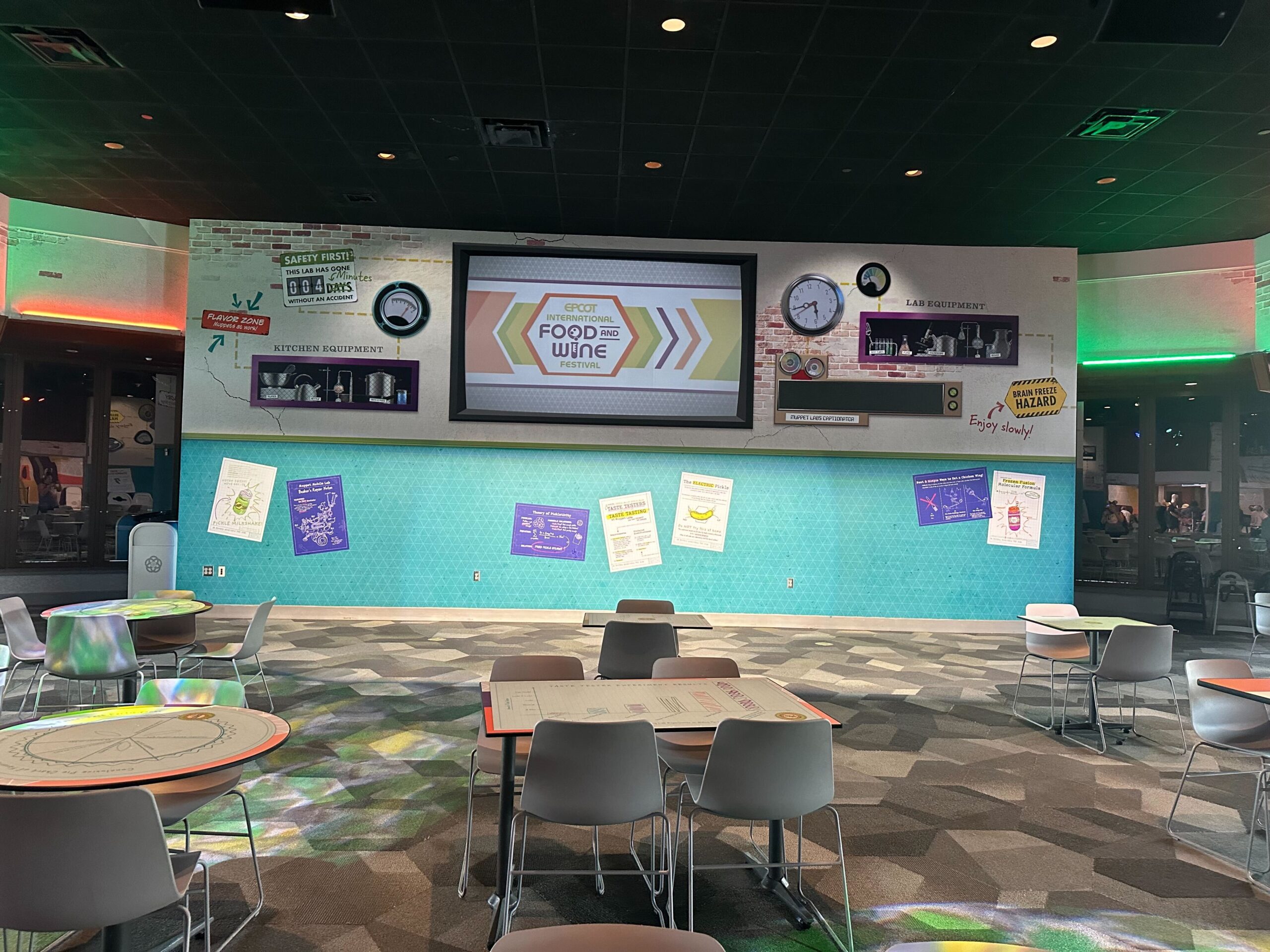 A spacious room features tables and chairs with a large screen showing "Epcot Food & Wine" on a wall decorated with various posters and design elements. Multicolored lighting accents the area.