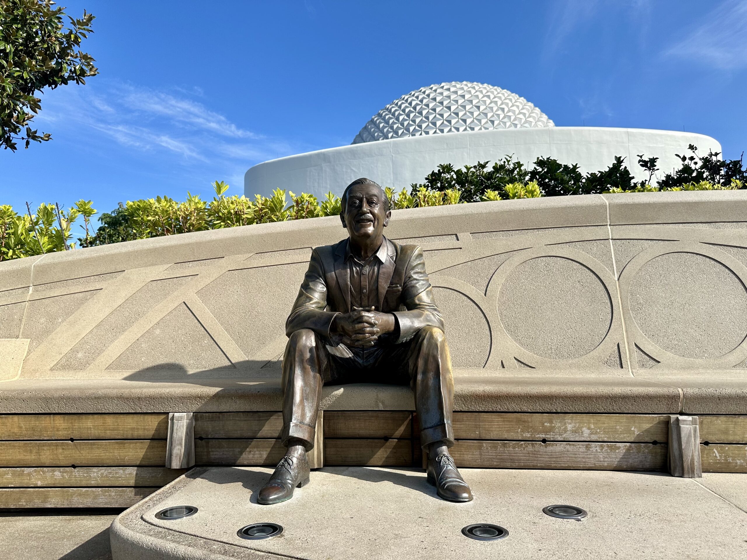Bronze statue of a man in a seated position, situated on a concrete structure with a geodesic dome in the background under a clear blue sky.