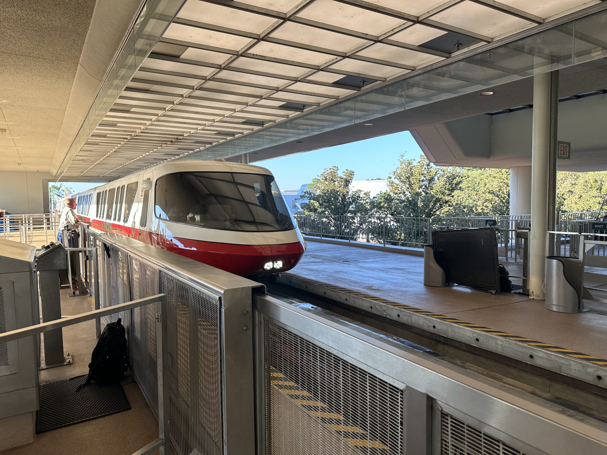 A red and white monorail train is parked at an elevated station platform. Trees are visible in the background under a clear sky.