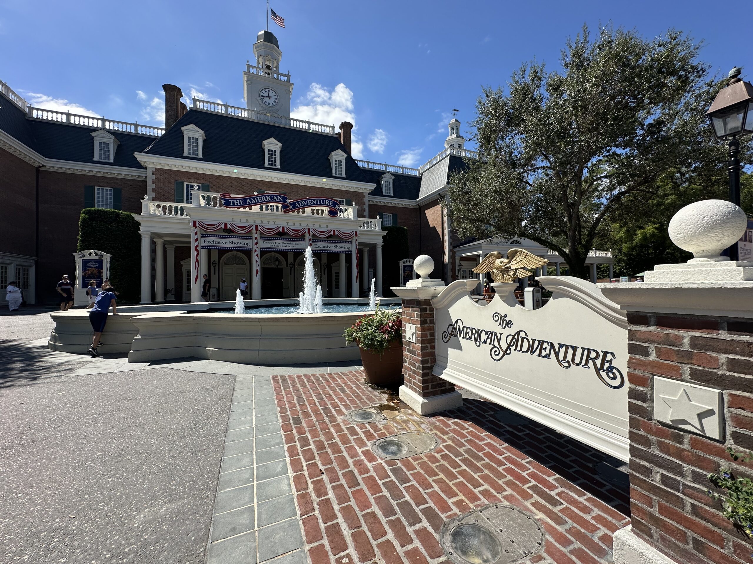 Historic building with a clock tower and American flag, featuring a brick pathway, a fountain, and a sign reading "The American Adventure" with decorative elements and a tree in the background.