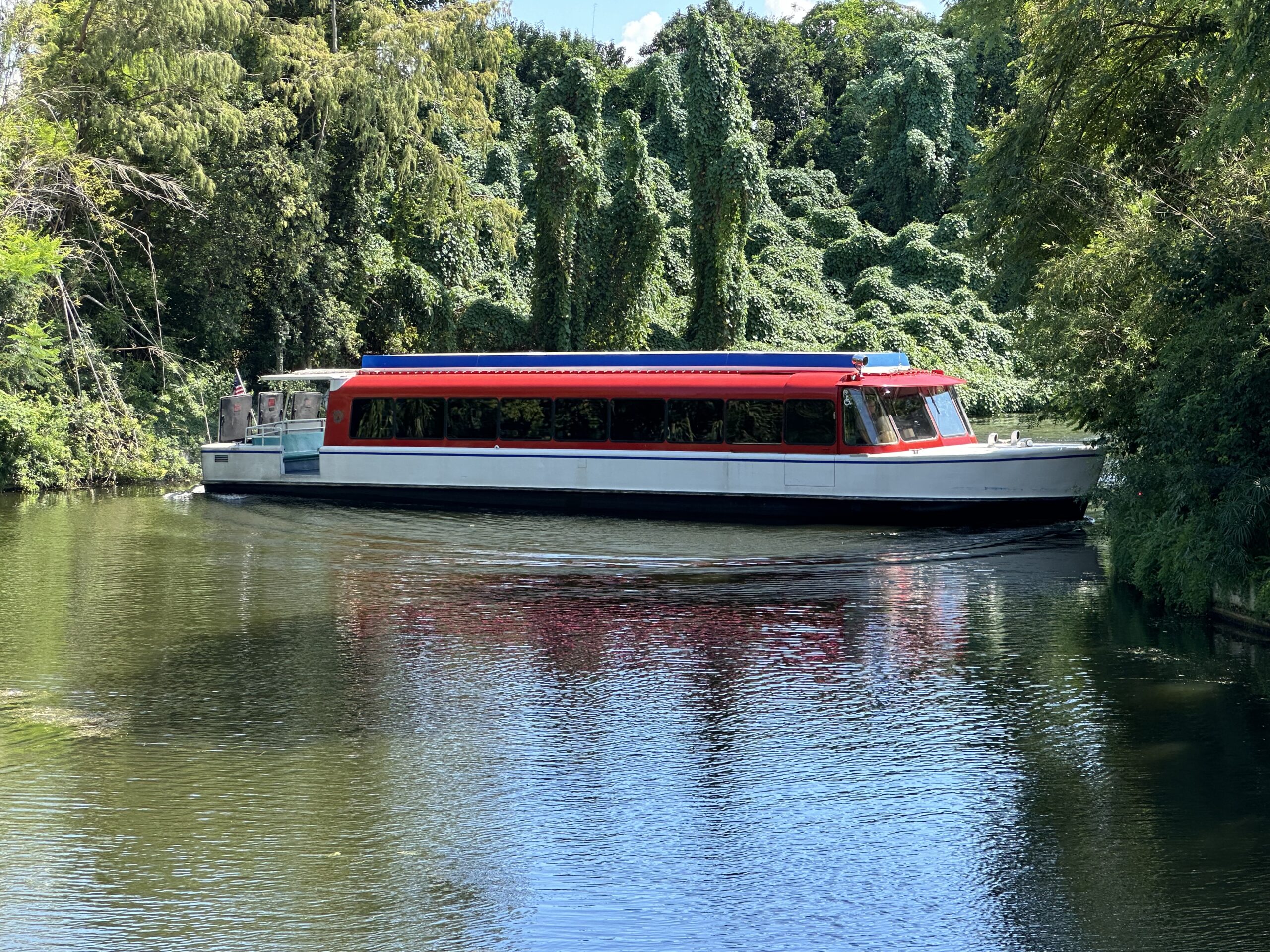 A red and white tourist boat navigates a serene, tree-lined river bend under a sunny sky.