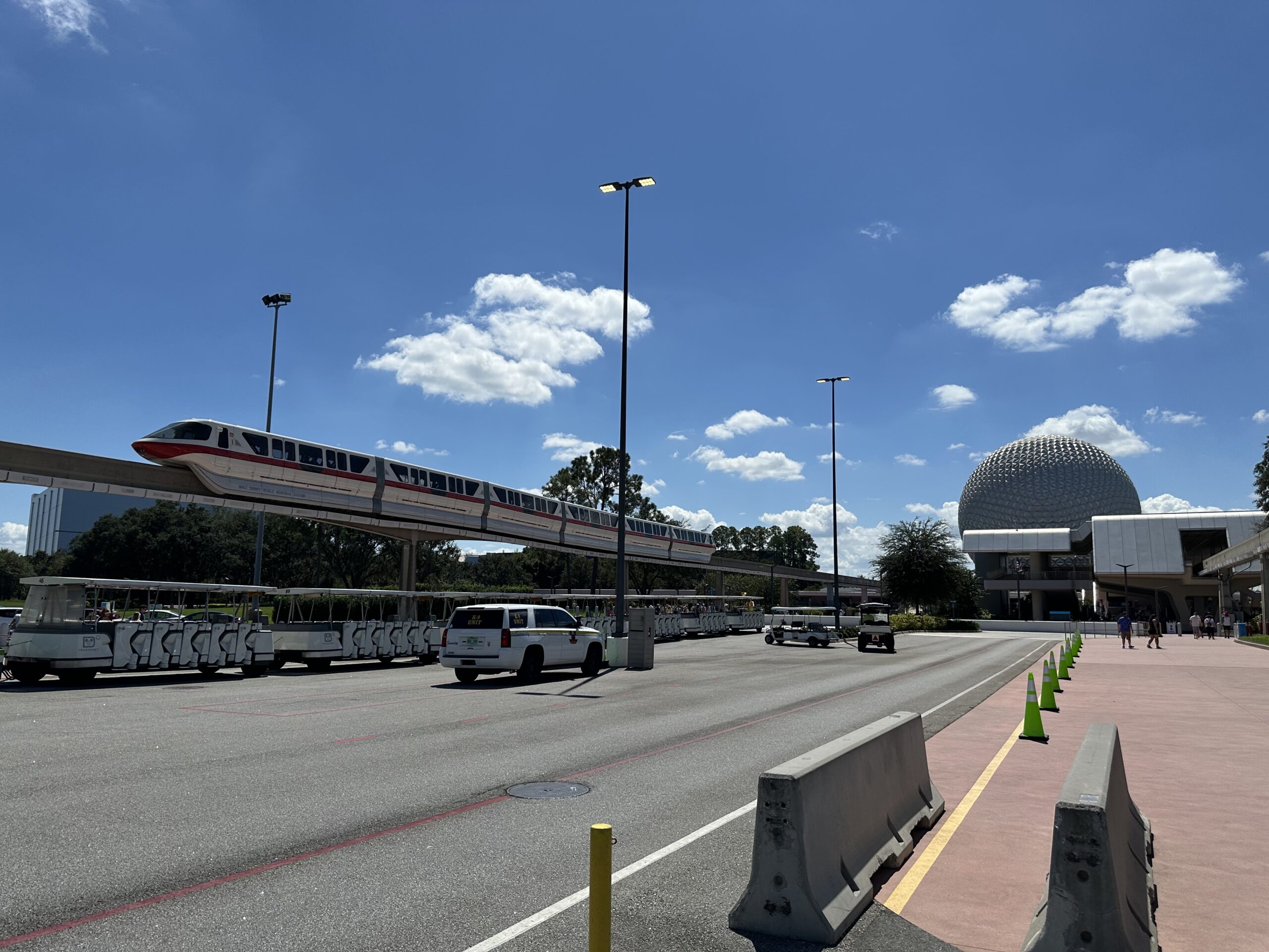 A monorail train travels on an elevated track near Epcot in Disney World, Florida, with Spaceship Earth visible in the background under a bright blue sky with scattered clouds.