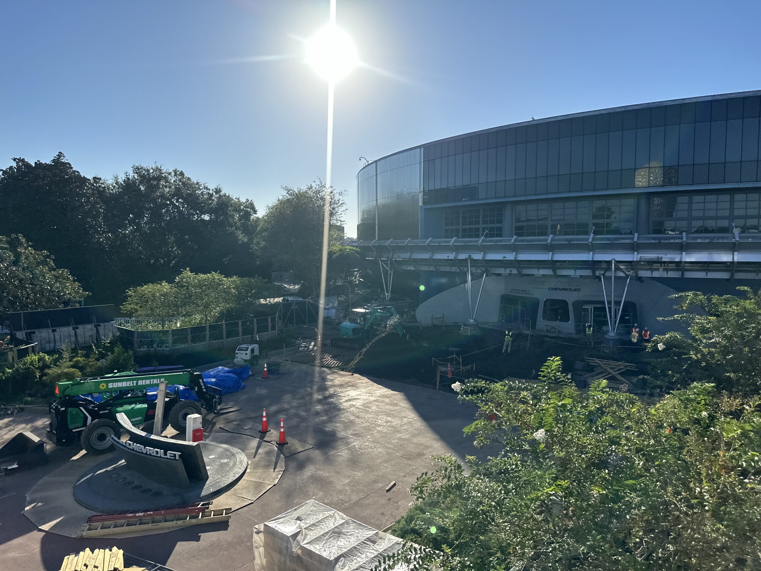 Large construction site with several workers, machinery, and materials in front of a modern glass building under clear sunny skies.
