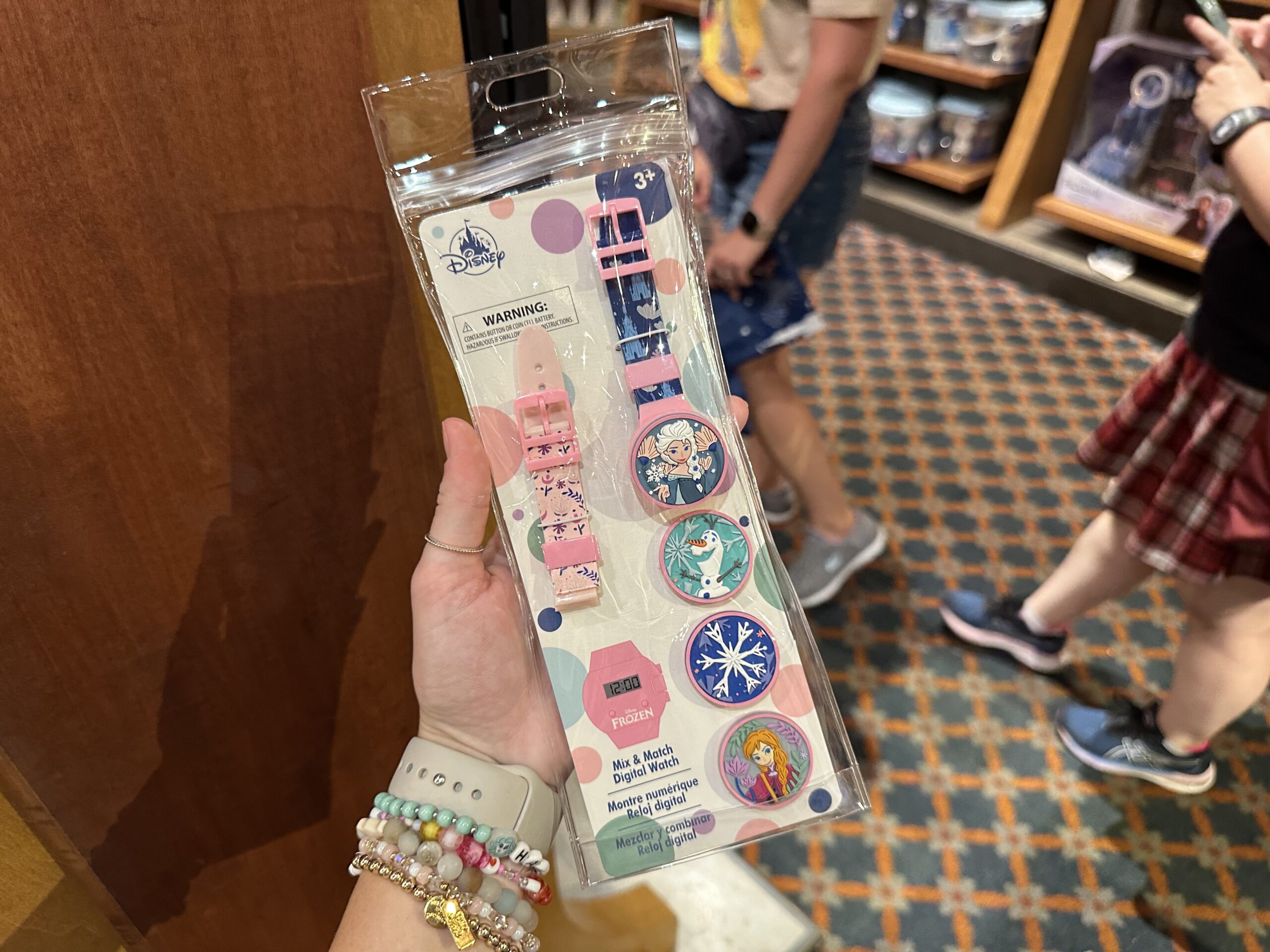A hand holds a packaged Disney-themed 3-piece Me & My Mini luggage accessory set, featuring various character designs. Background shows people in casual attire standing on a checkered carpet.