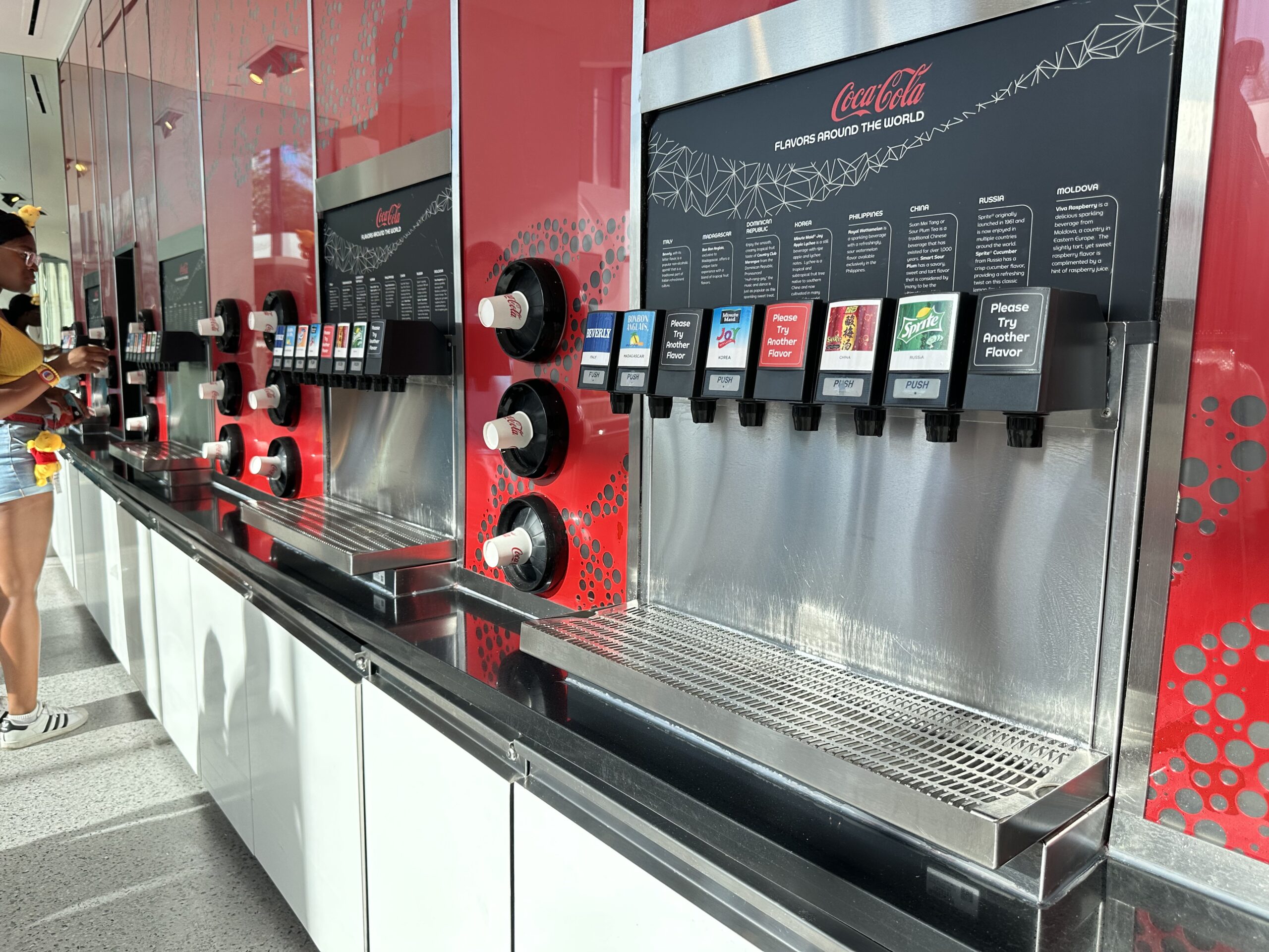 A row of Coca-Cola soda dispensers, offering various flavors from around the world, installed on a red and white wall in a self-service area.