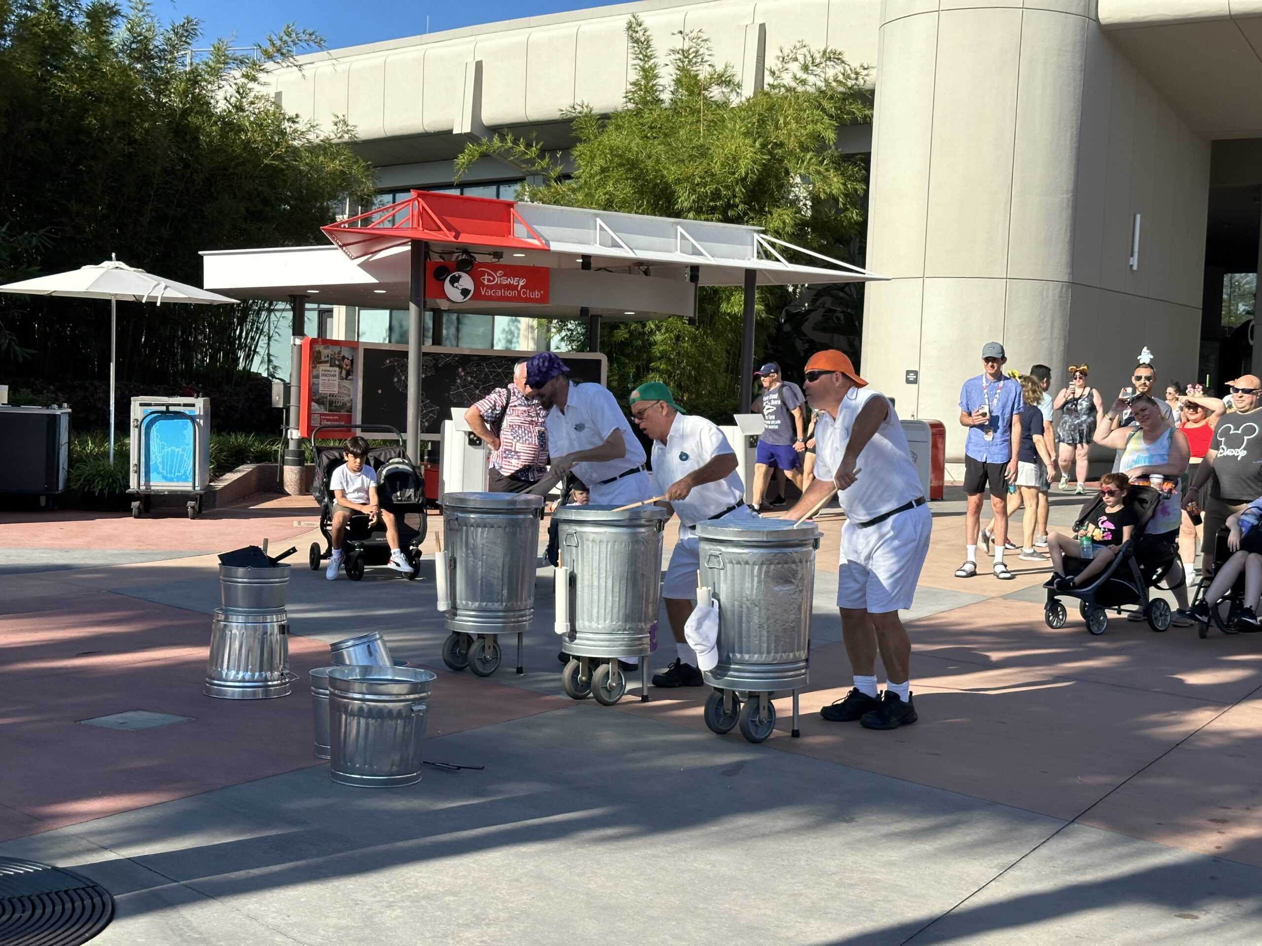 Three performers dressed in white uniforms play music on metal trash cans as a small crowd watches in an outdoor plaza.