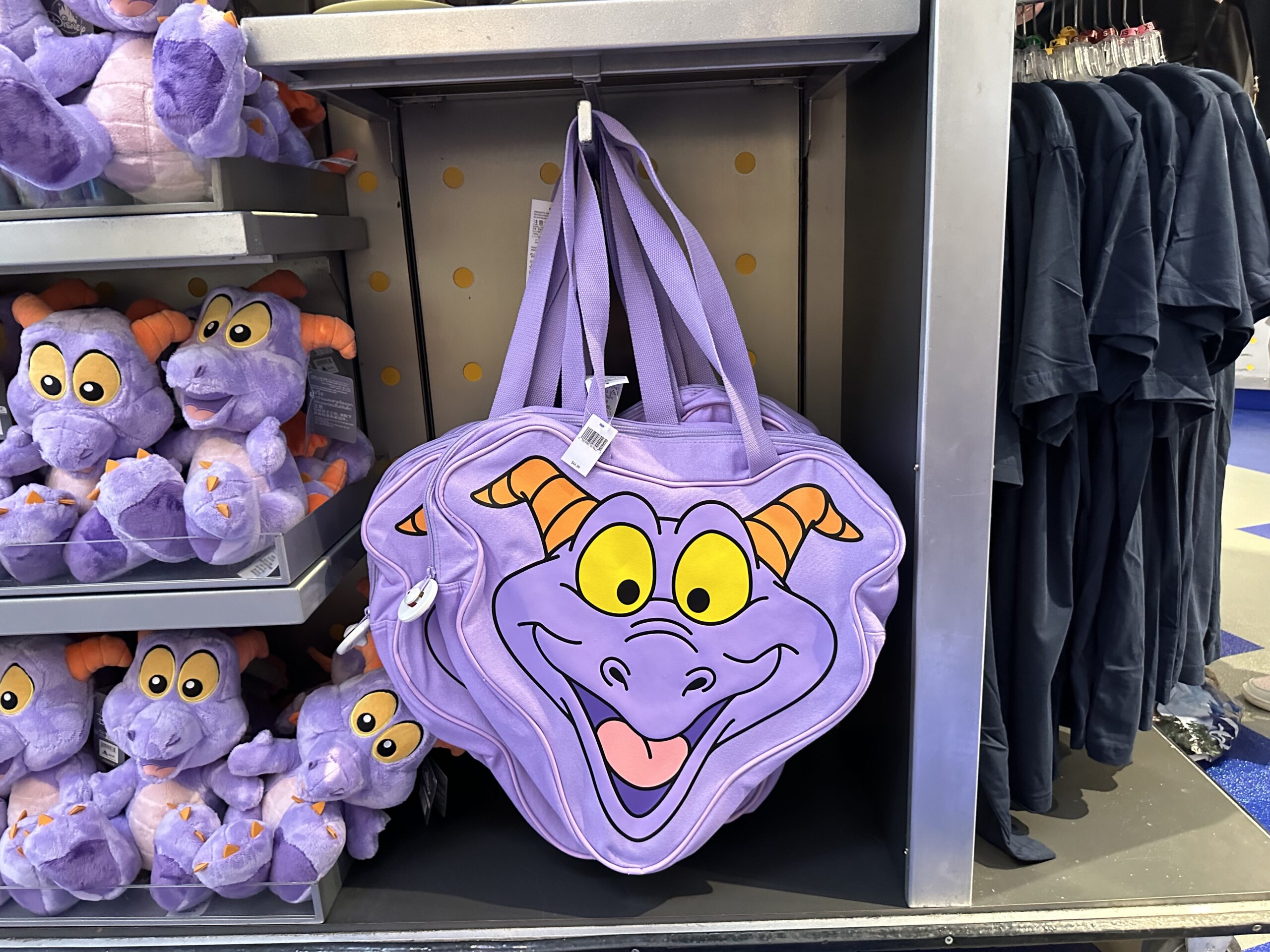 A shelf display featuring plush toys and a large purple bag with a cartoon dragon face. The bag has yellow eyes, orange horns, and a wide smile.