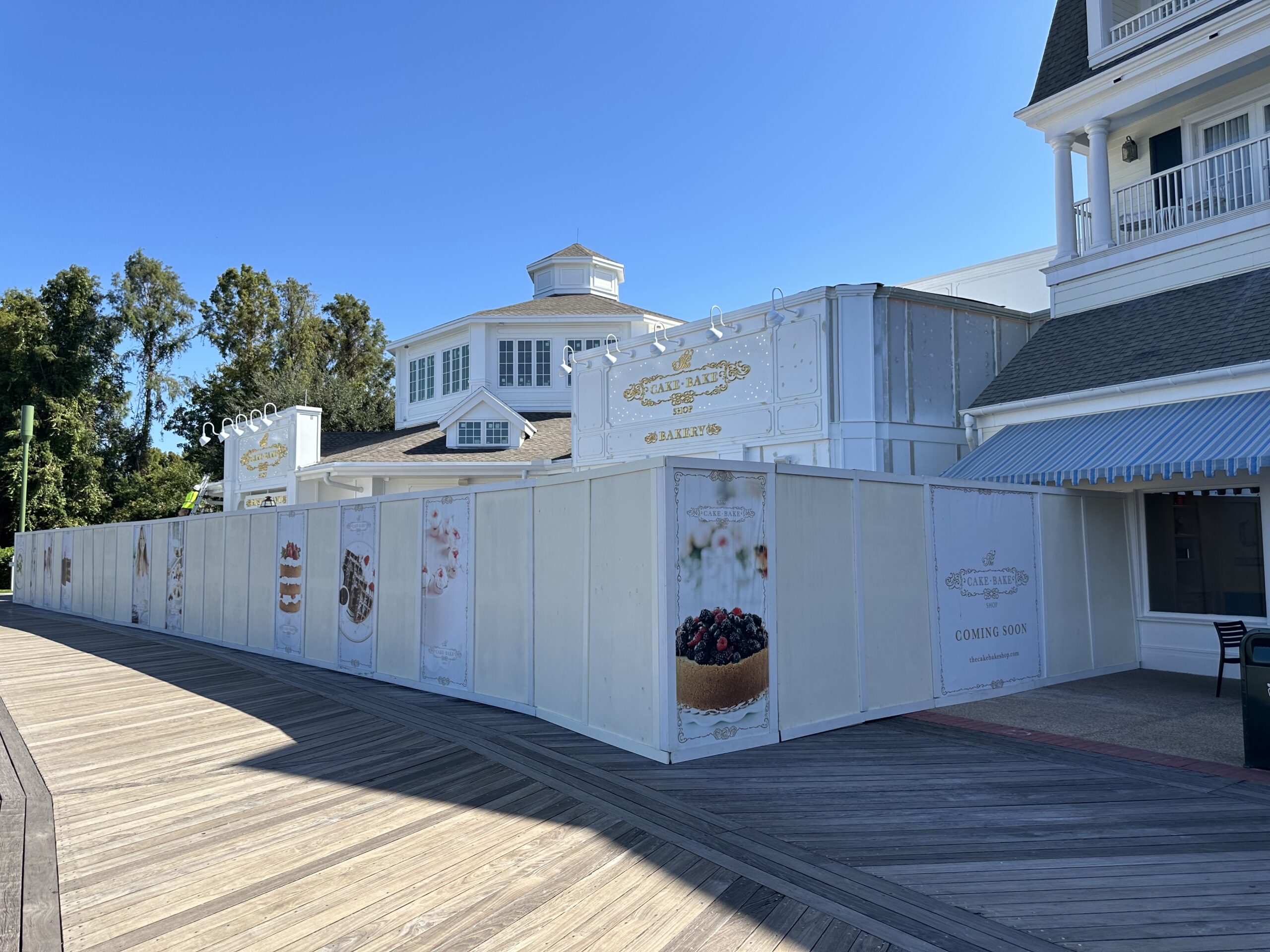 Construction site of a future bakery, surrounded by white barriers with dessert images and "Coming Soon" signs, located along a wooden boardwalk under a clear blue sky.