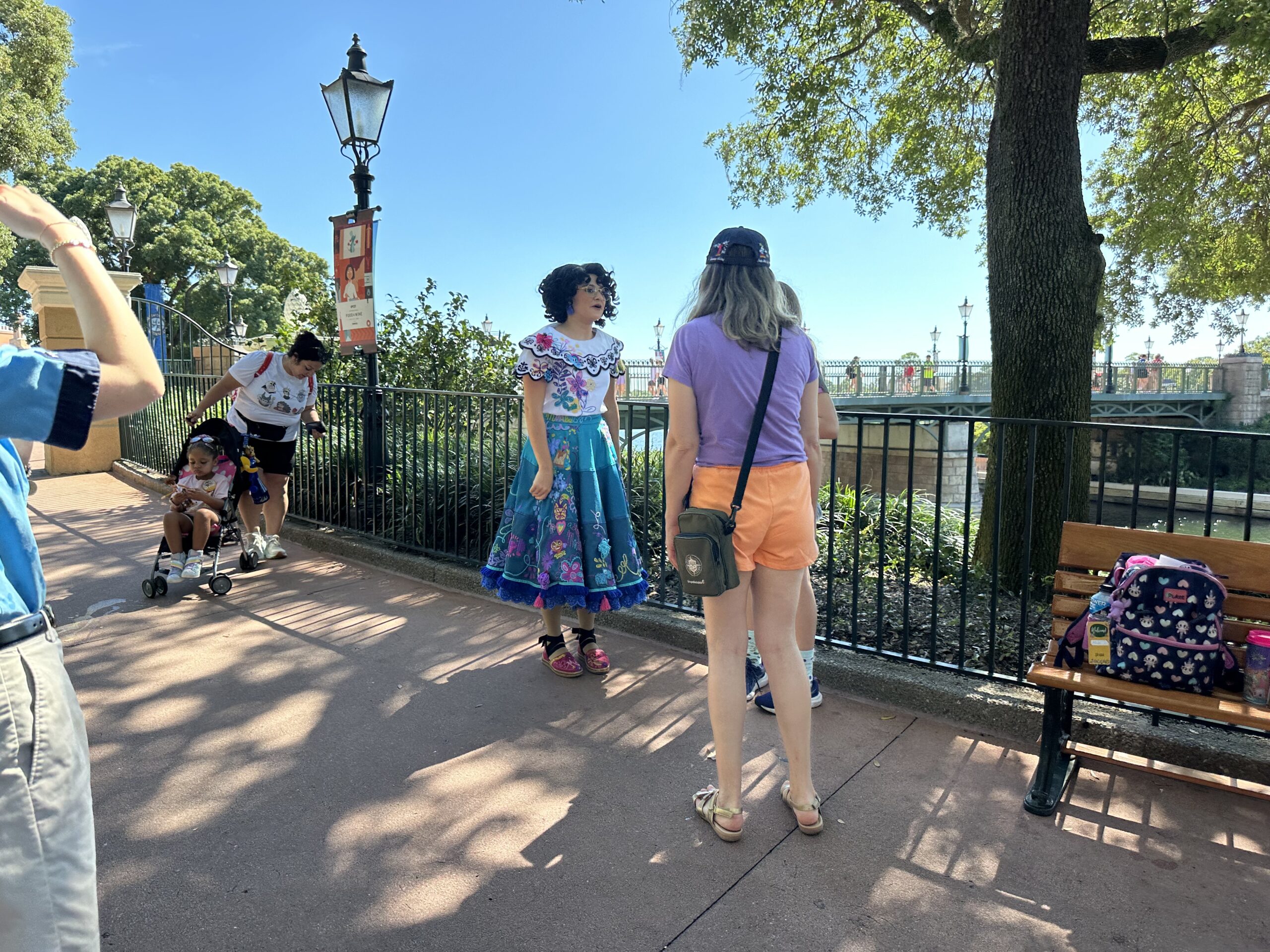 Two people are chatting in an outdoor setting near a bench and a lamppost. One person is dressed in a colorful costume, and another is wearing casual clothing. A stroller with a child is nearby.