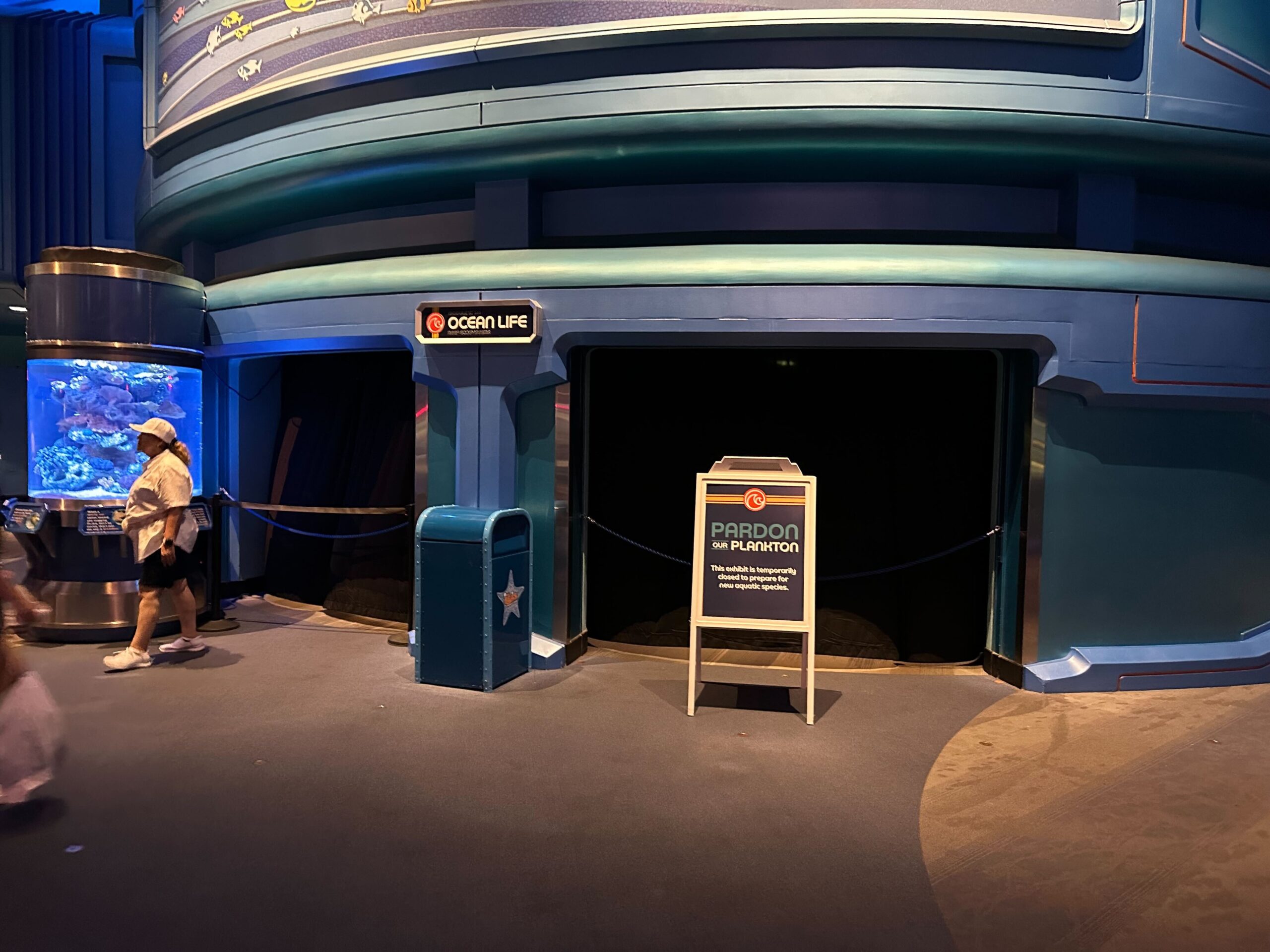 A blue-themed aquarium display with a "Pardon the Plankton" sign and black curtains covering the entrance. A person walks by on the left, past a trash bin and an aquarium tank that echoes the mysteries of the seas.