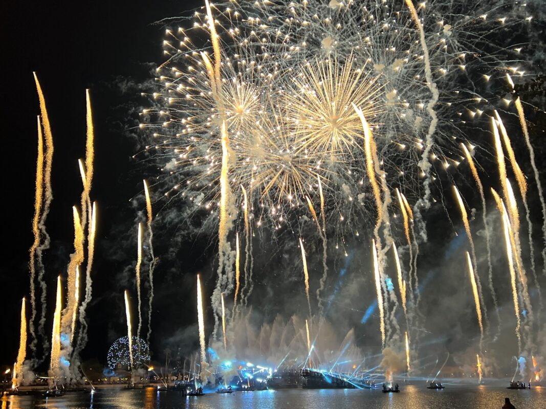 A nighttime display of fireworks with numerous bright streaks and large bursts illuminating the sky over a large body of water. A ferris wheel is visible in the background to the left.