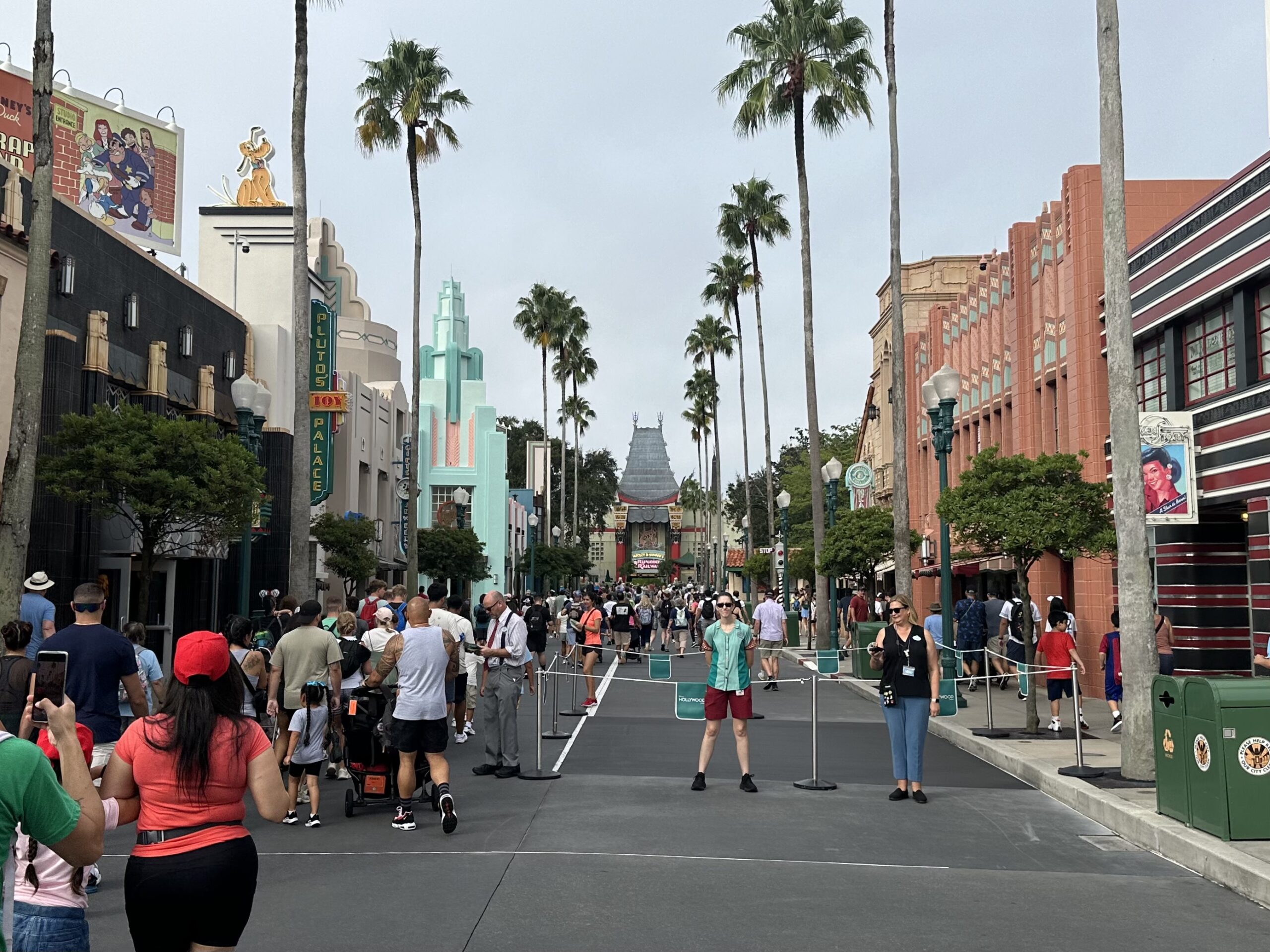 Hollywood Studios entrance past the touchpoints reveal a bustling street with people walking, palm trees lining the road, and an ornate building, replica of the Chinese Theater in California, in the background under a gray sky. Rope barriers block freshly poured concrete.
