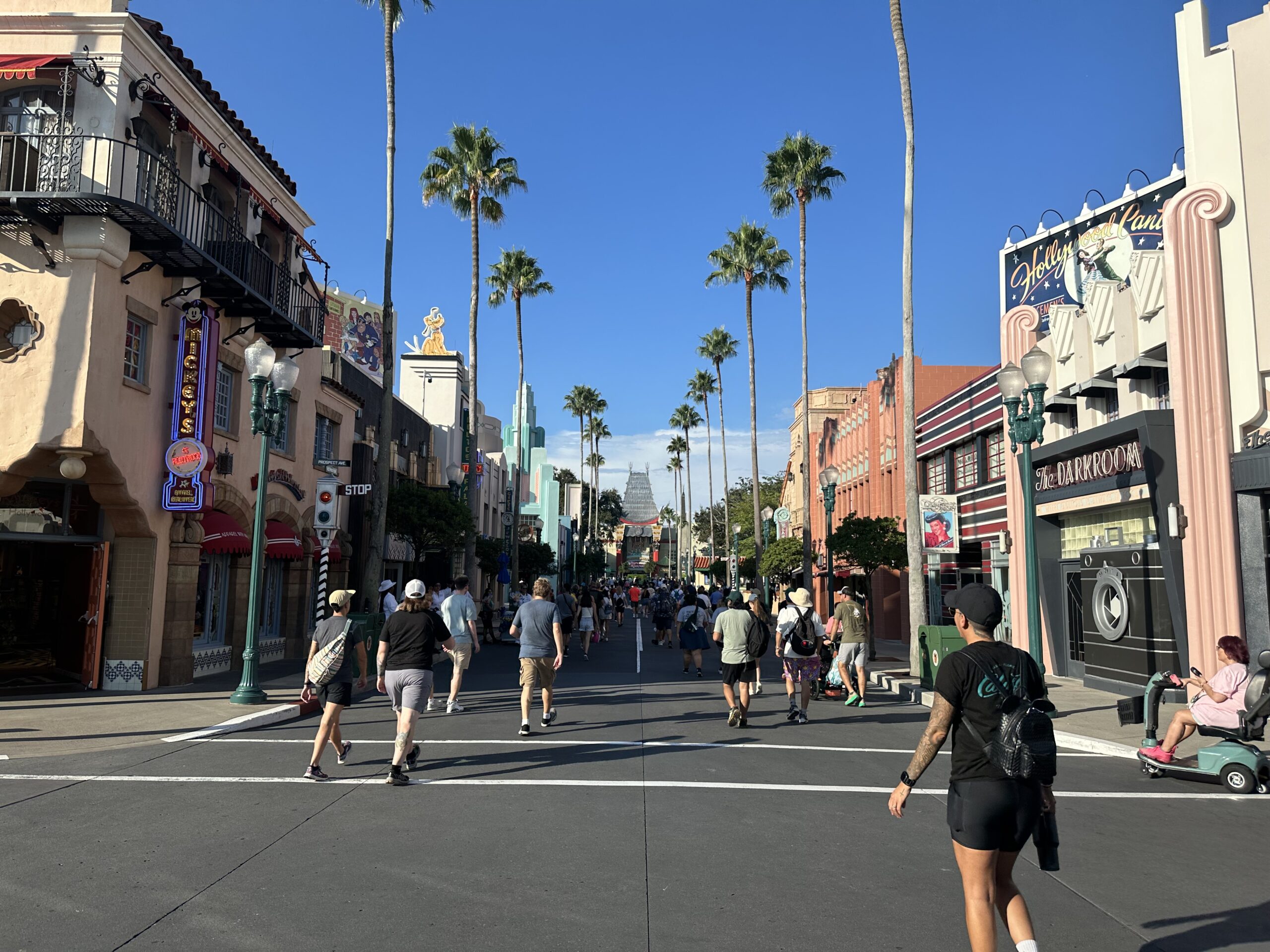 A crowd of people are walking on a main street lined with palm trees and buildings on a sunny day. Signs for shops and eateries are visible on either side. Hollywood Studios