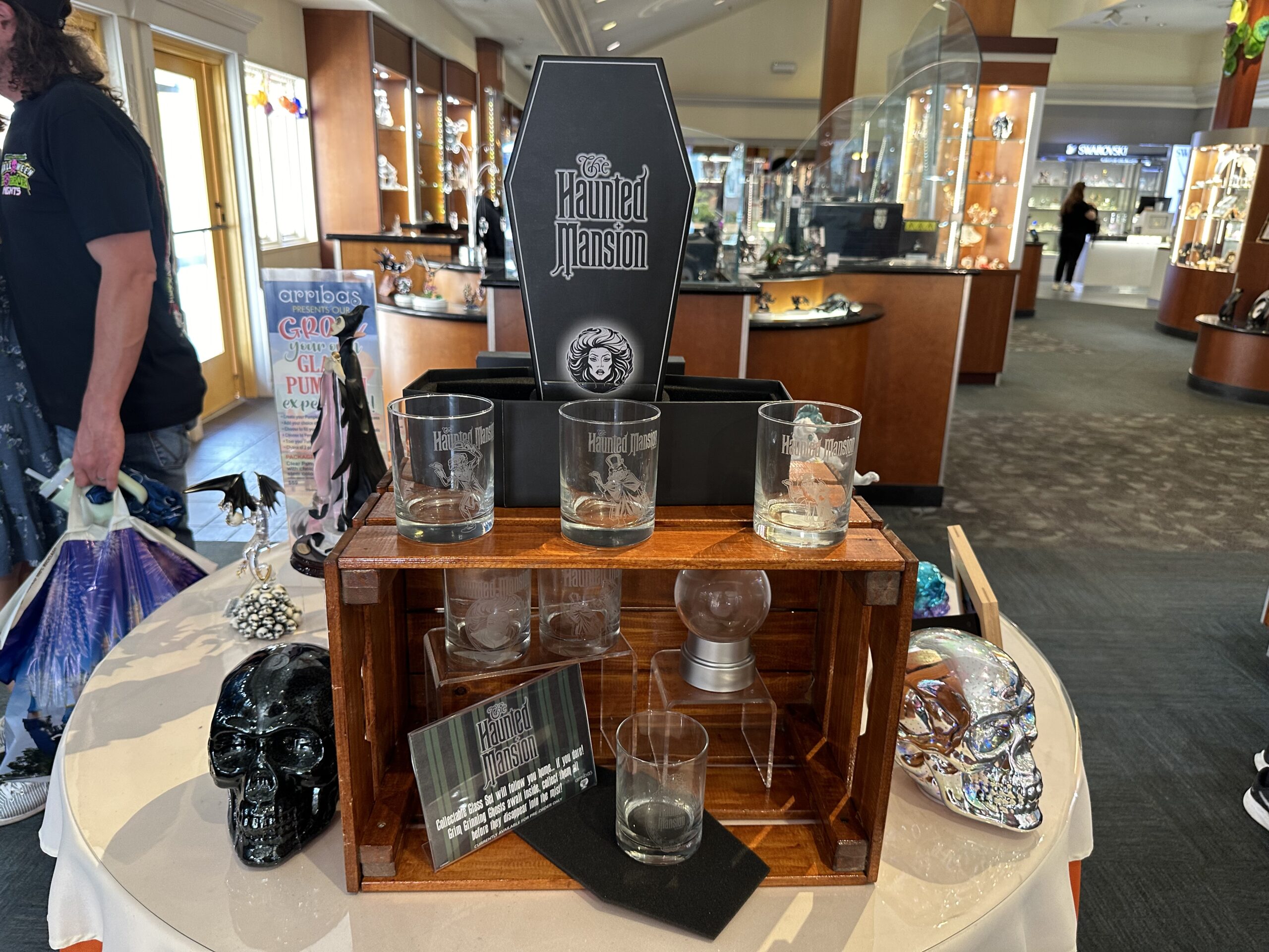 A display table showcasing "The Haunted Mansion" themed glassware, skull decorations, and a coffin-shaped sign inside a store.