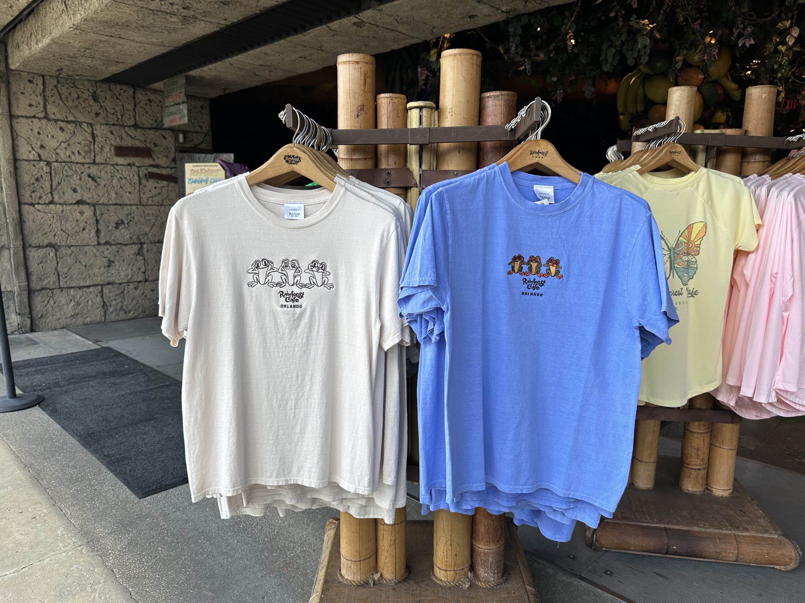 Display of various colored t-shirts on wooden stands, including beige, blue, and yellow options, arranged outside a storefront with stone walls.