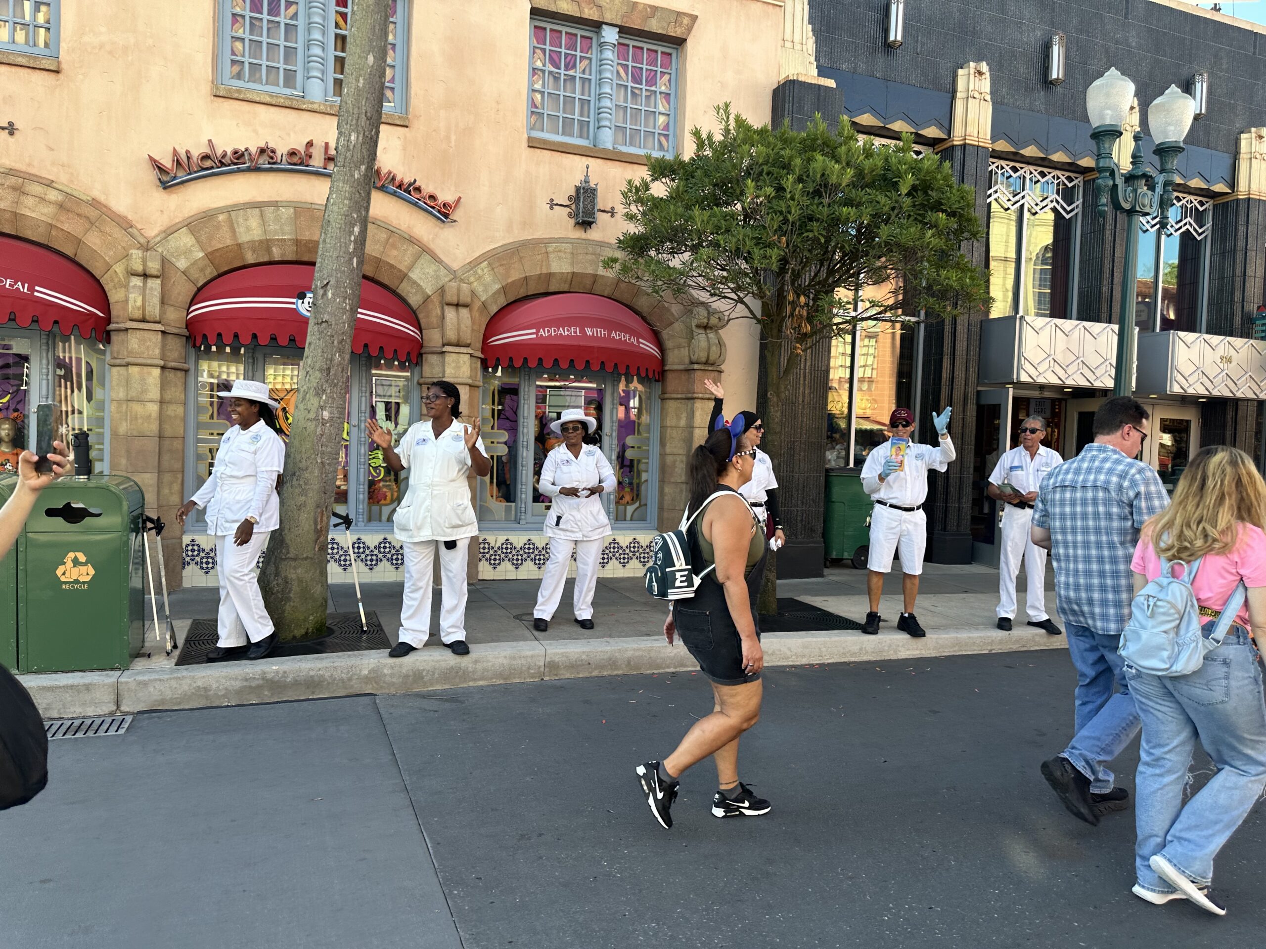 A group of performers in white uniforms stand in front of a storefront, interacting with passing visitors on a busy street.