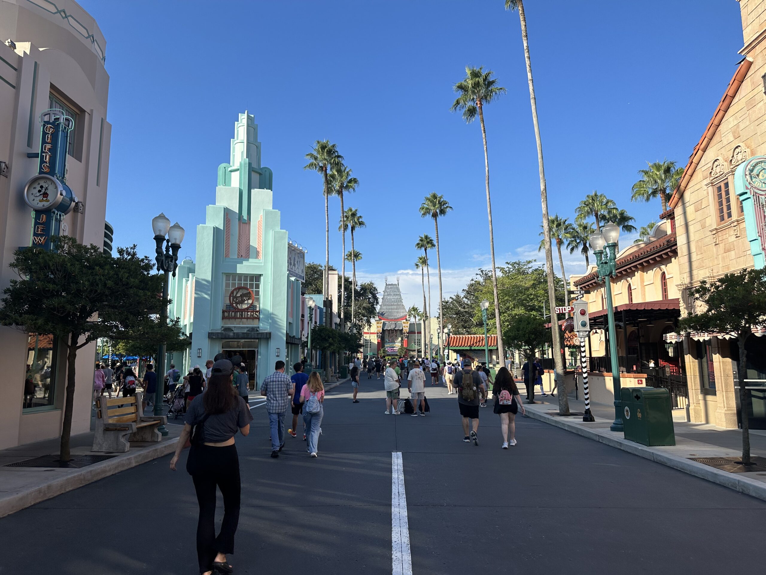 People are walking down a wide, palm tree-lined street in a theme park, surrounded by retro-styled buildings under a clear blue sky. Hollywood Studios