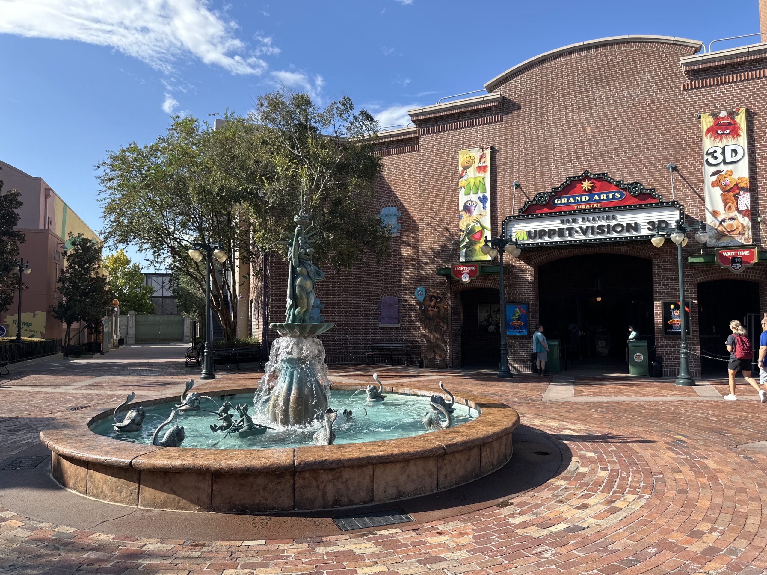 A circular fountain with statues in a courtyard in front of a brick theater building with a sign for "Muppet Vision 3D." Two vertical banners on the building show images of Muppet characters.