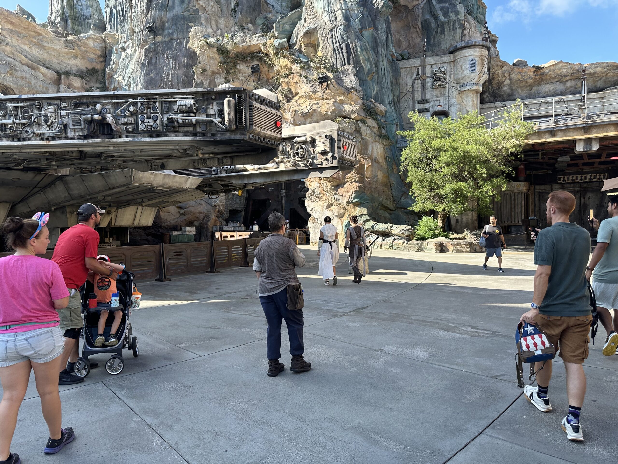 Visitors walk through an outdoor area with rocky terrain and themed structures, including a spacecraft. Some people push strollers, and others carry backpacks.
