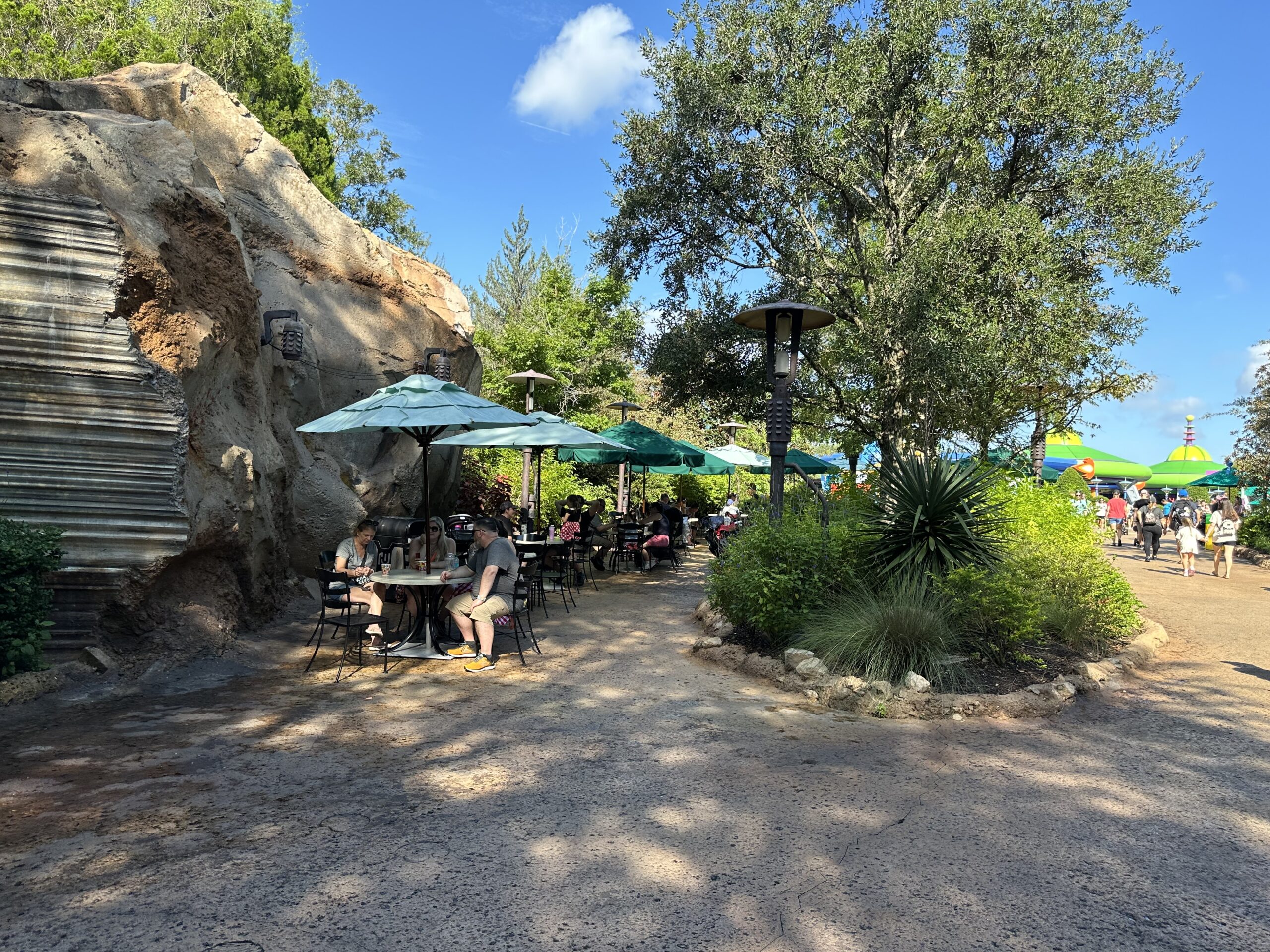 People are dining at outdoor tables with green umbrellas near a rocky outcrop and trees on a sunny day.