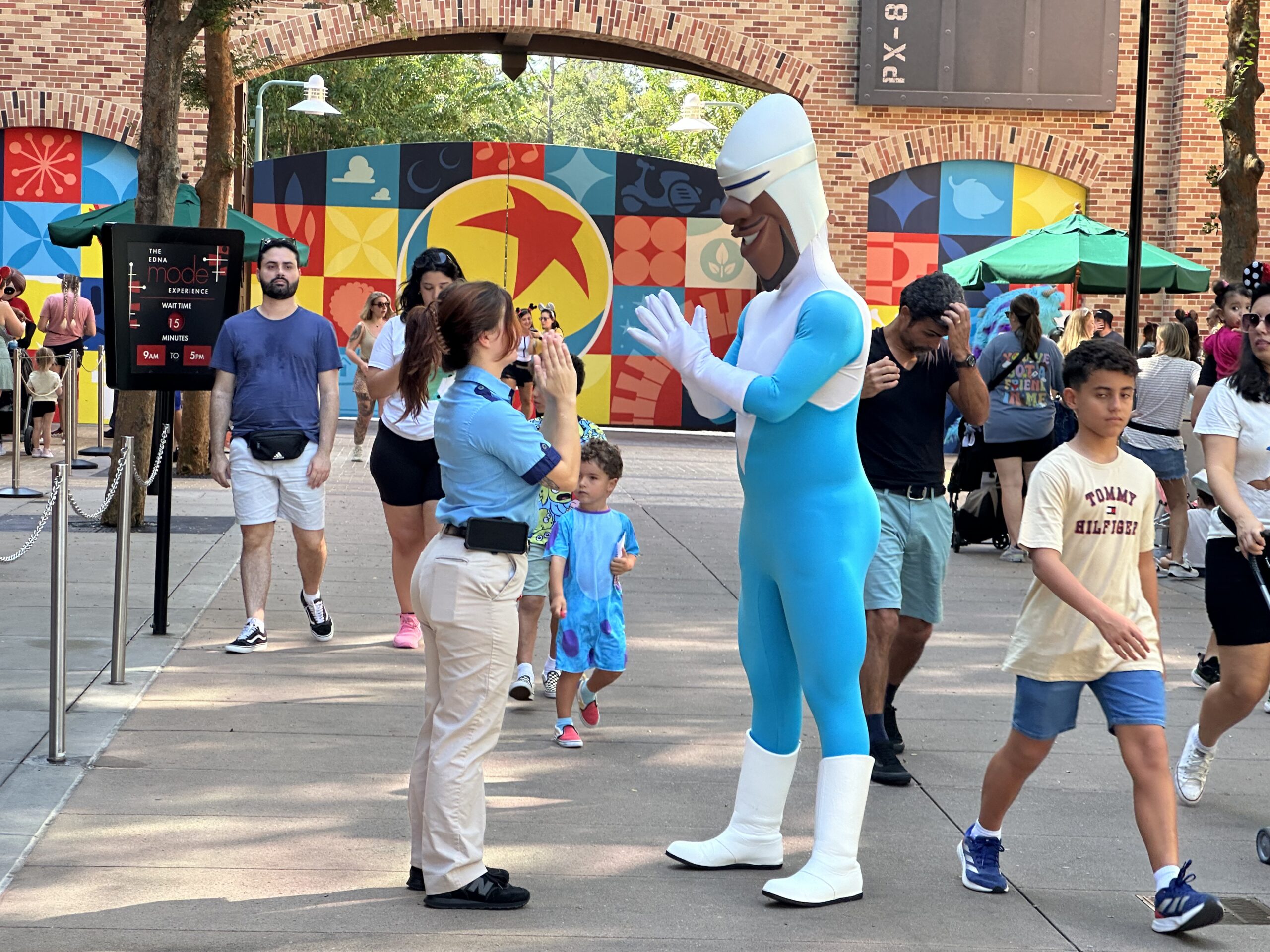 A person in a blue and white superhero costume interacts with a woman and a young child at an outdoor venue. Other visitors and colorful wall art are visible in the background.