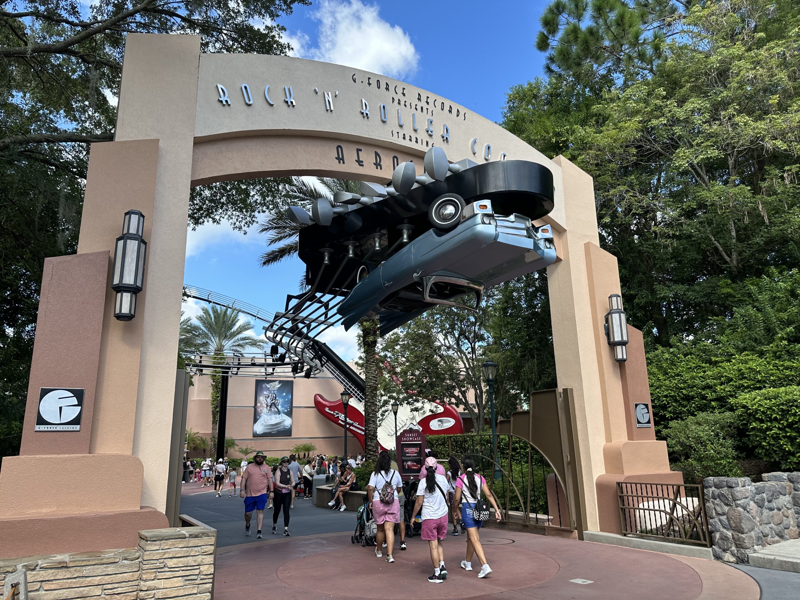 People walk under a large archway with a suspended car at the entrance of a theme park attraction. Trees and a roller coaster are visible in the background.