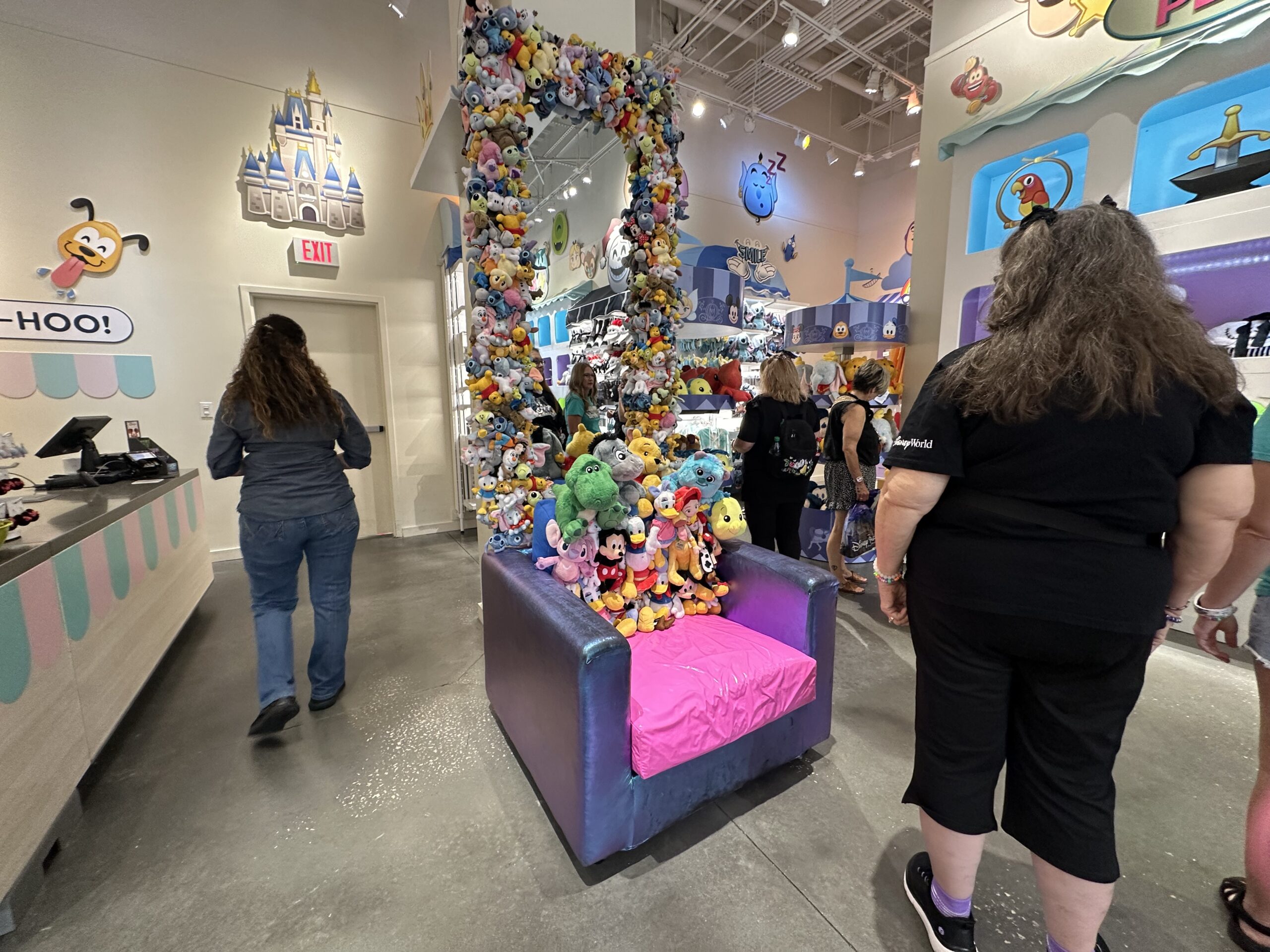 A store interior displays a large chair covered in various stuffed toys, surrounded by more plush toys hanging in an arch above it. People browse the store.
