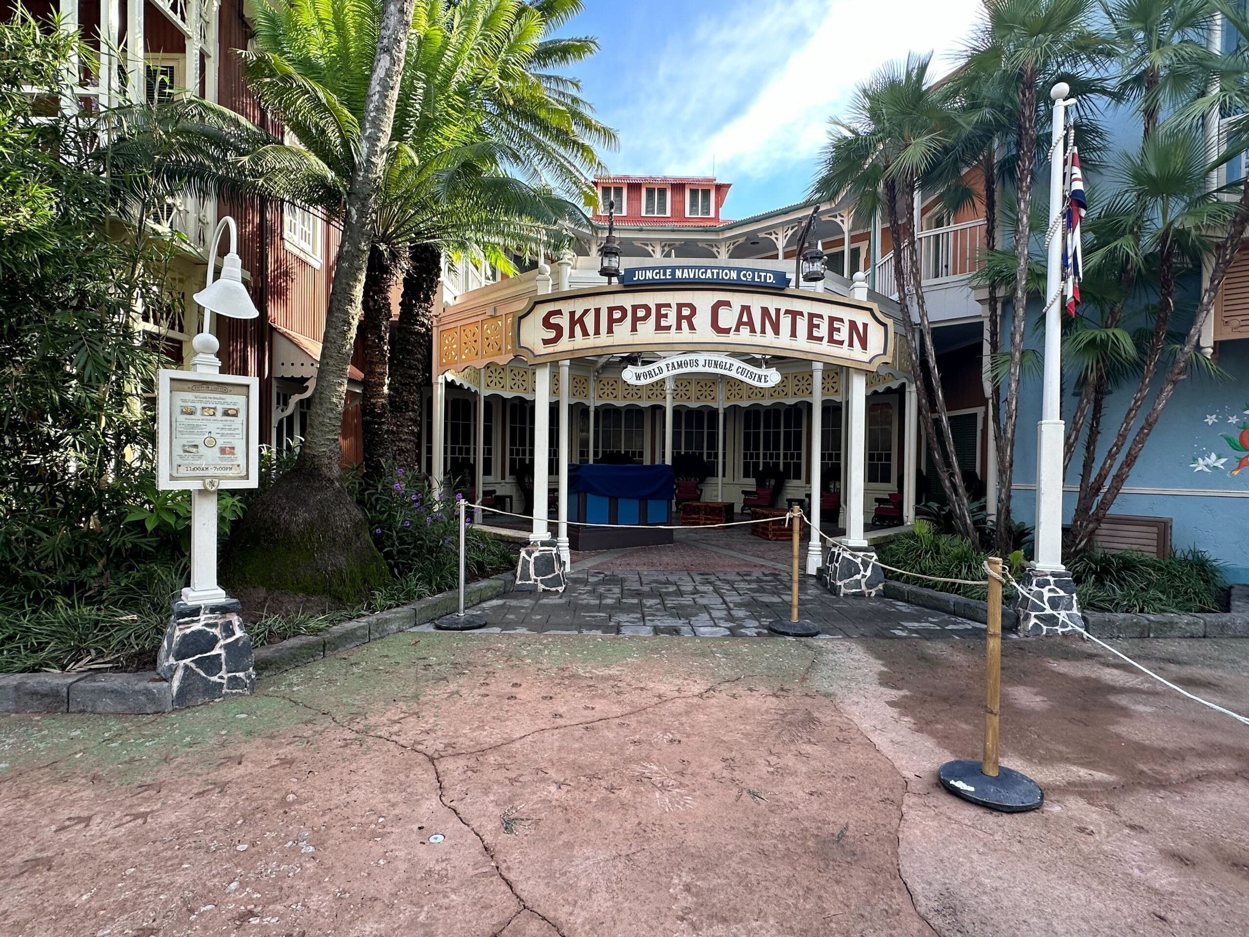 Entrance of Skipper Canteen, a restaurant with an adventure theme, featuring a white and yellow sign, surrounded by tropical plants and rope barriers in an amusement park setting.