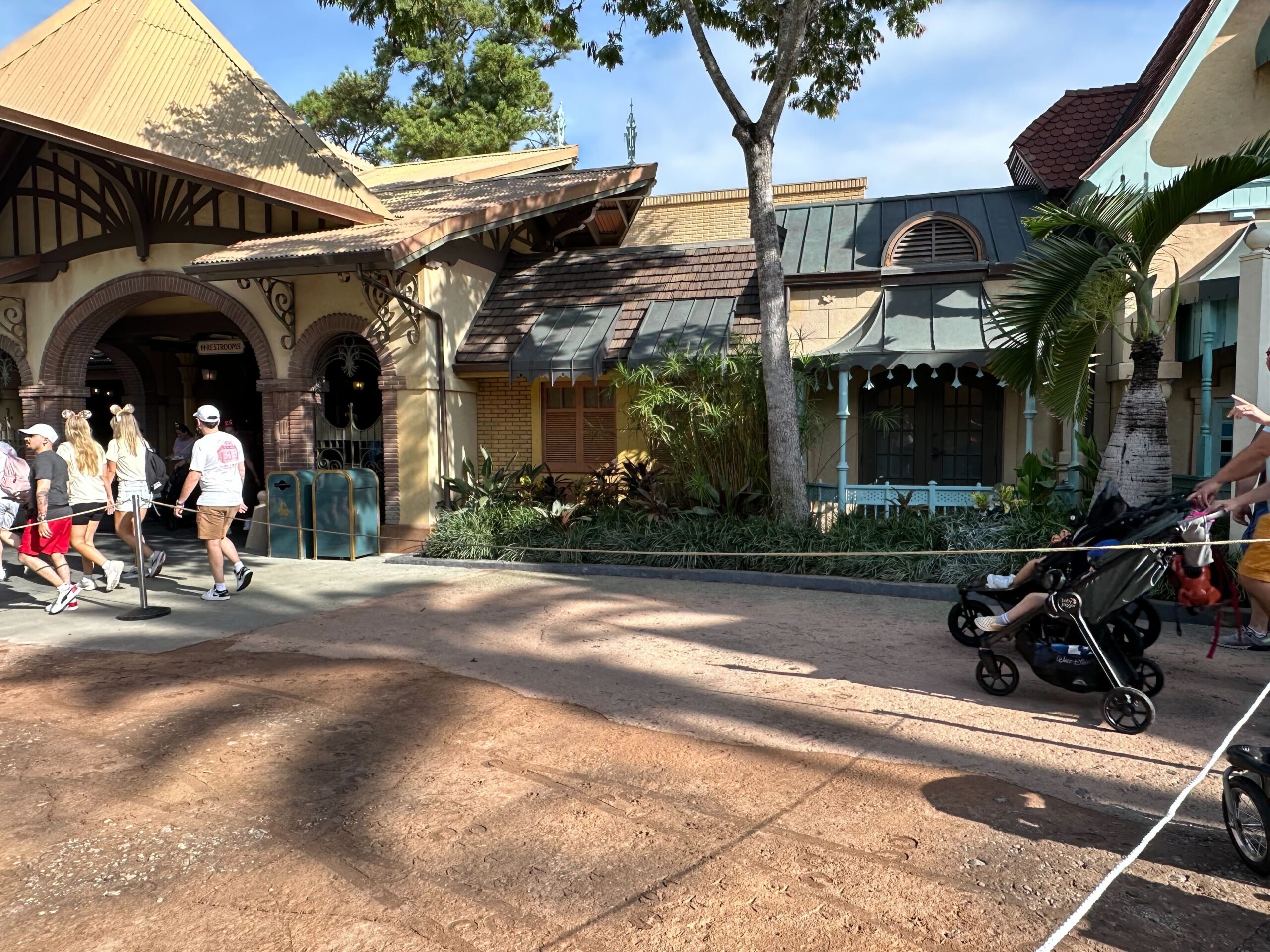 People walk by an entrance to a building on a sunny day. A person pushes a stroller on the right side near a rope barrier. Trees and shaded areas are visible in the background.