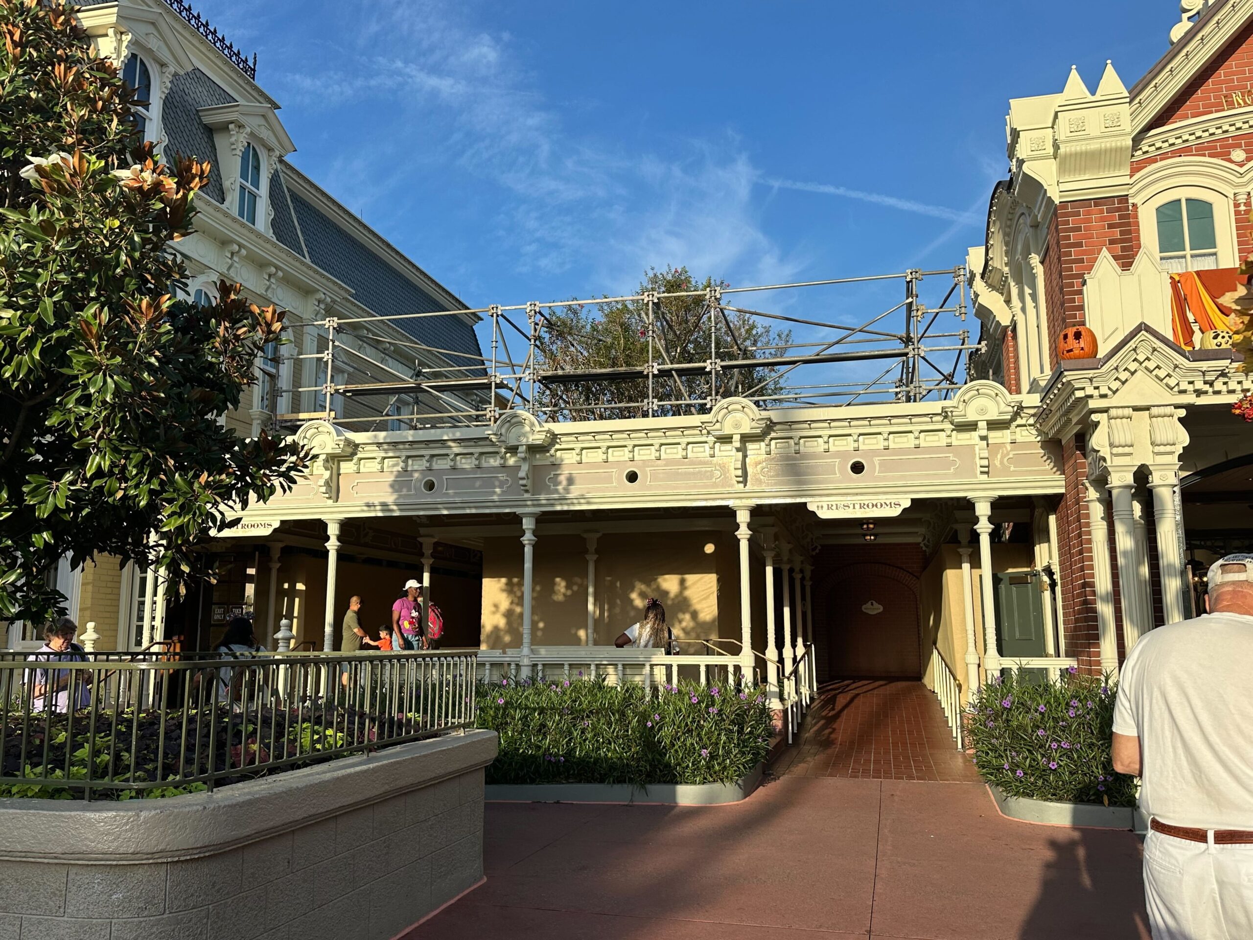 A building under renovation with scaffolding on its facade, located between two other structures. A few people are visible near the entrance, and another person is in the foreground on the right.